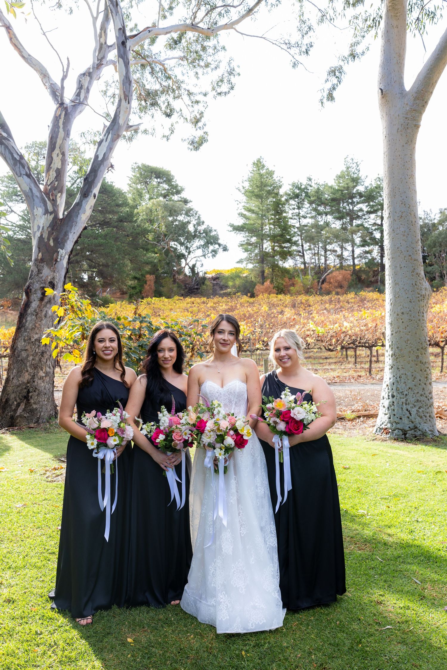Bride in white gown poses with three bridesmaids in black dresses holding colorful bouquets at vineyard wedding.