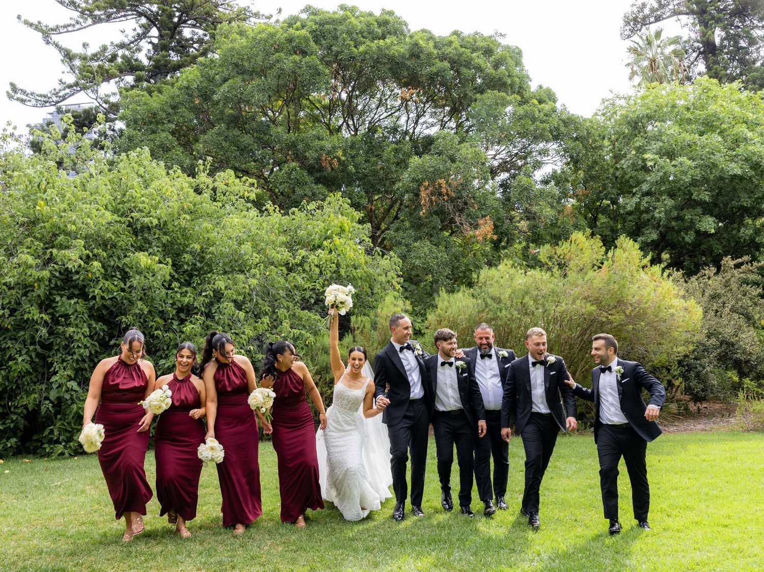 Wedding party in burgundy and black attire celebrating on a garden lawn.