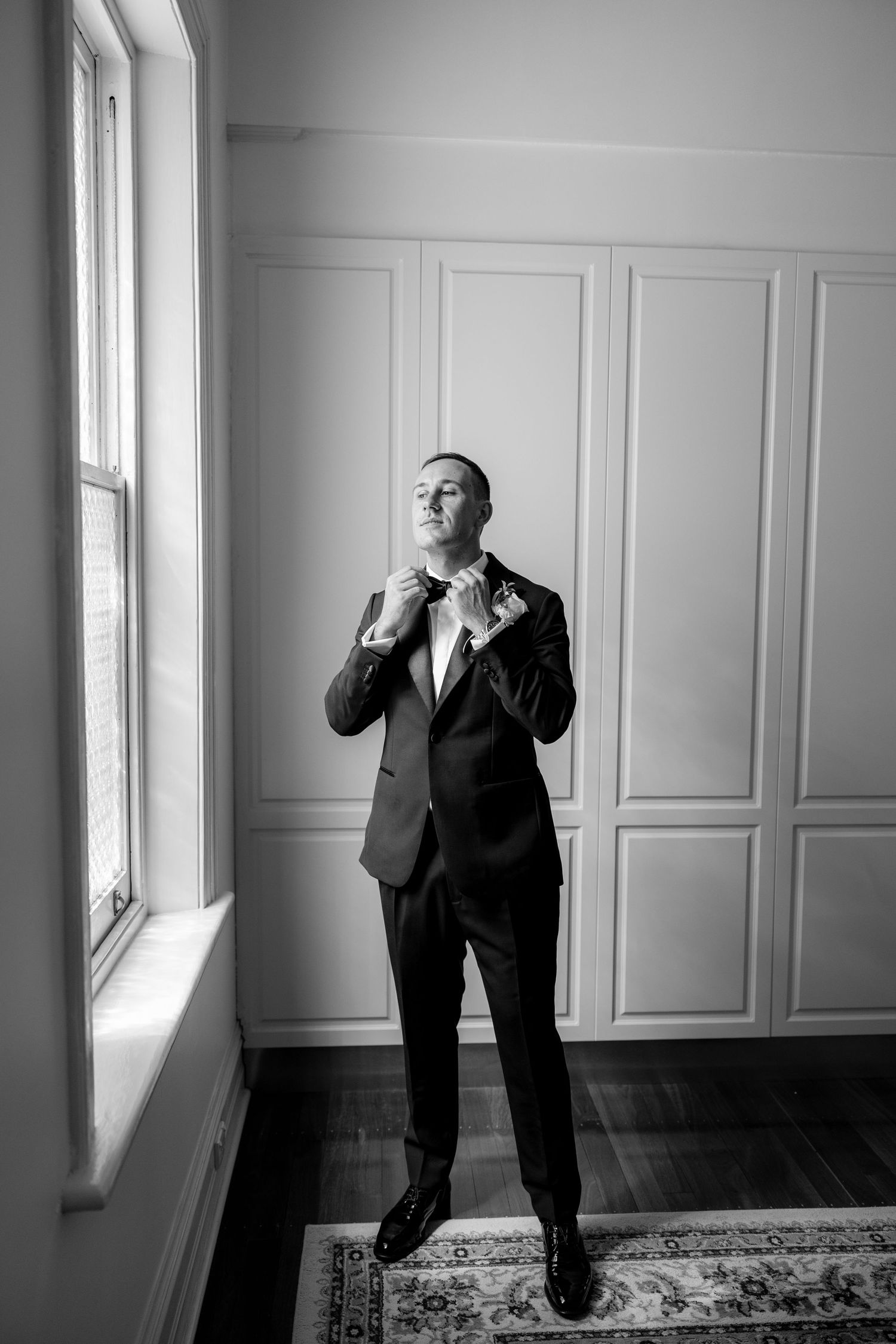 Groom in black tuxedo adjusts bow tie by window in black and white portrait.