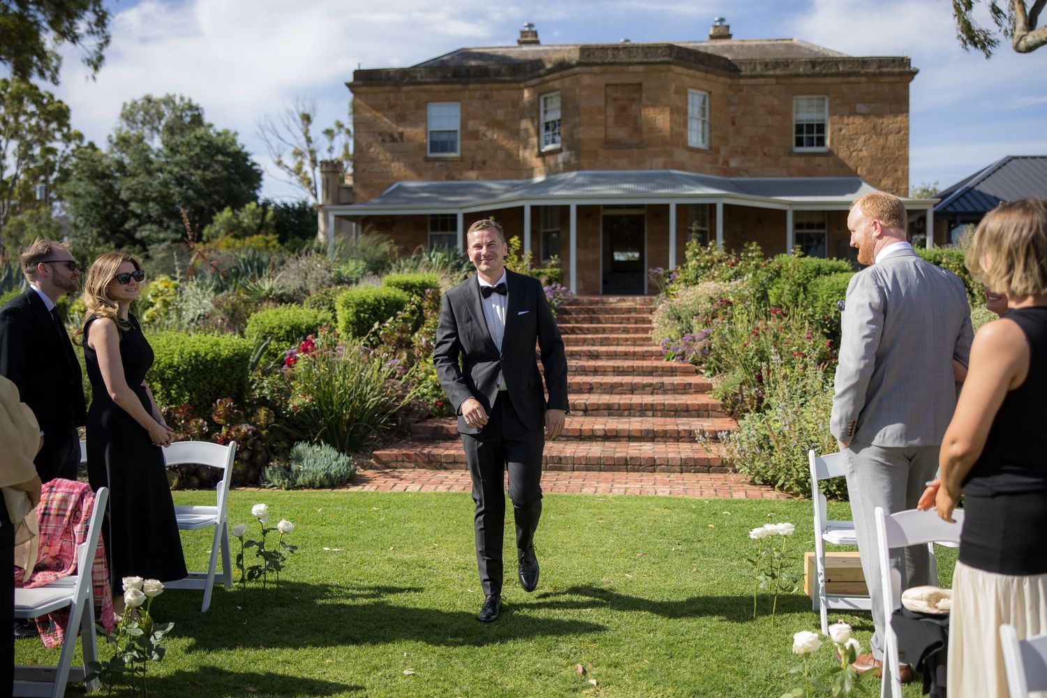 Groom walks down outdoor aisle toward altar at historic stone manor wedding venue.