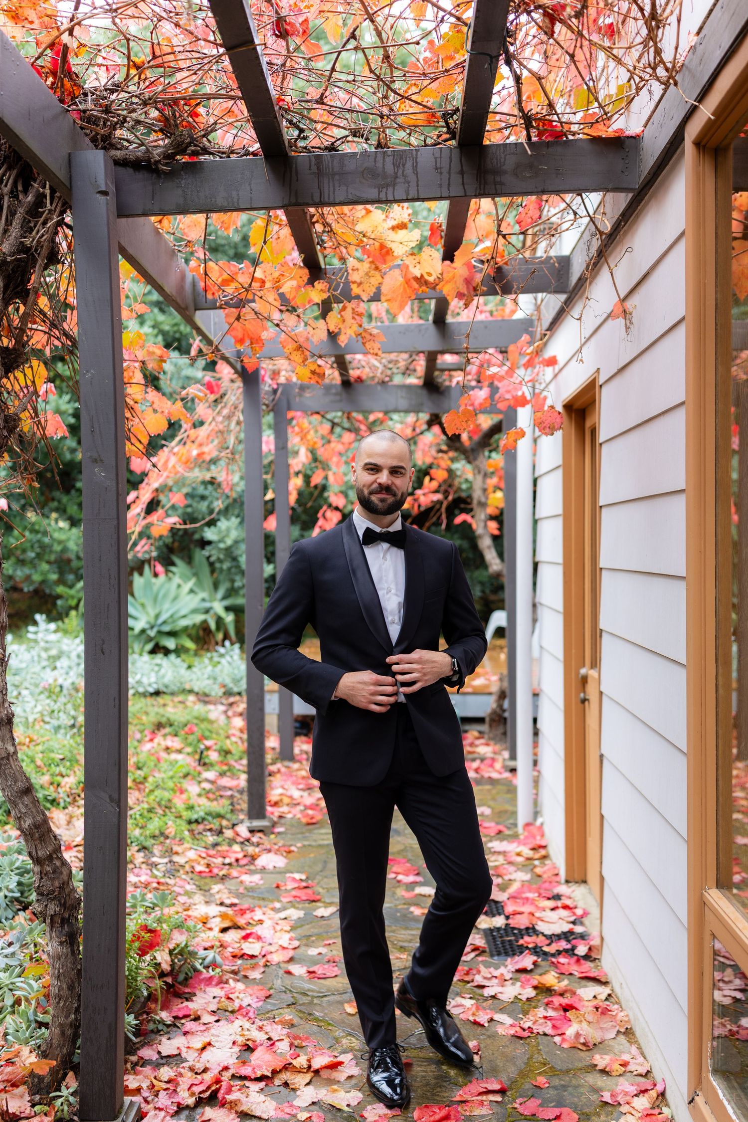 Dapper groom in black tuxedo with bow tie poses under pergola covered in brilliant red and orange autumn leaves.