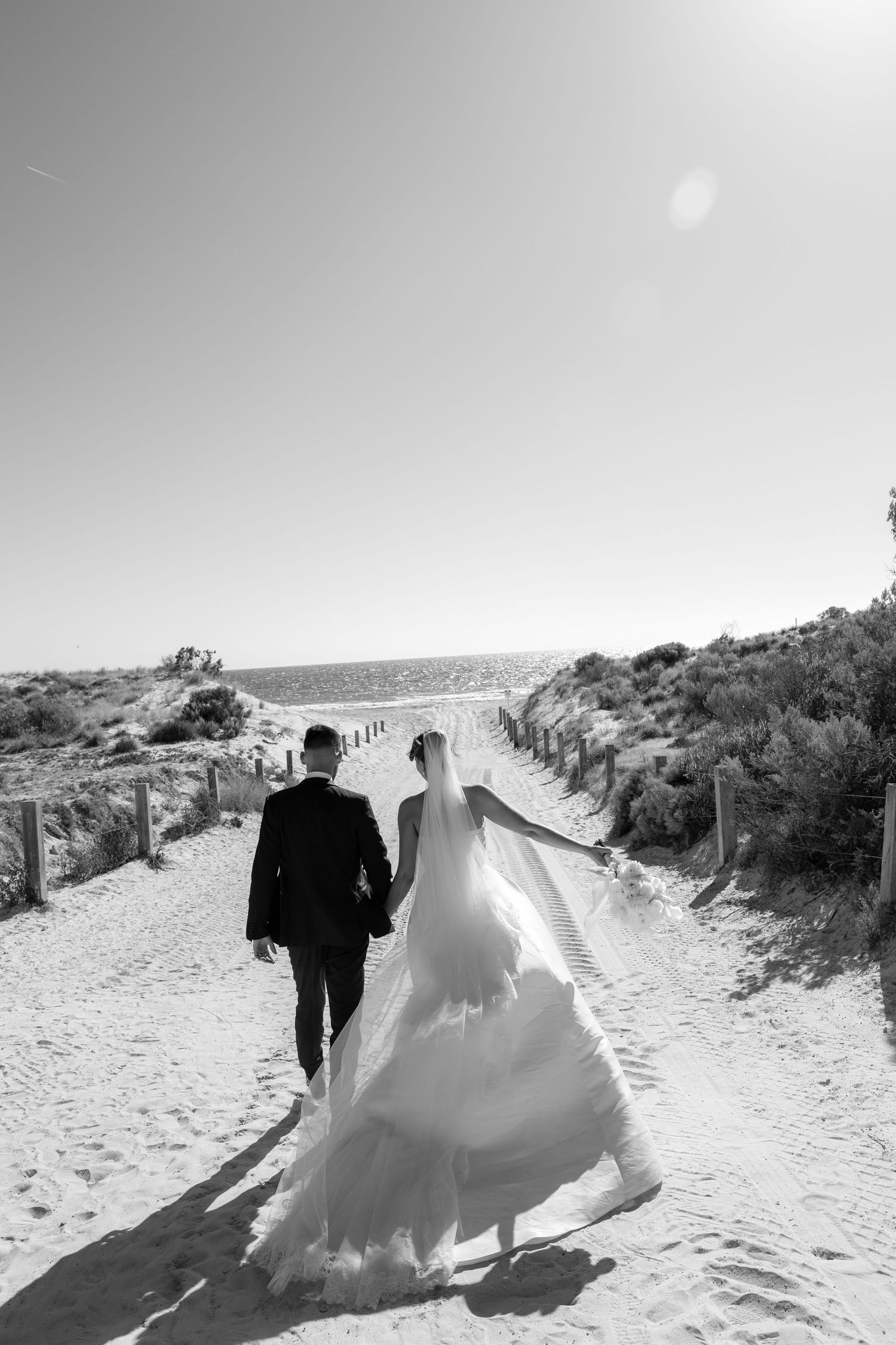 Black and white photo of a bride and groom walking hand in hand down a sandy beach path toward the ocean.
