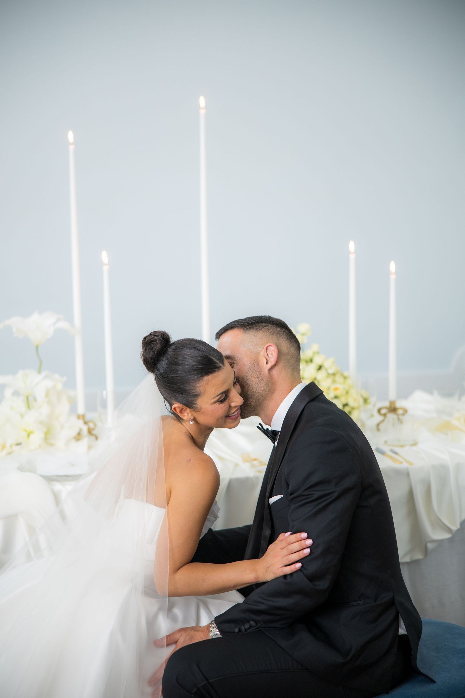 Bride and groom sharing intimate moment at decorated sweetheart table with tall white candles.