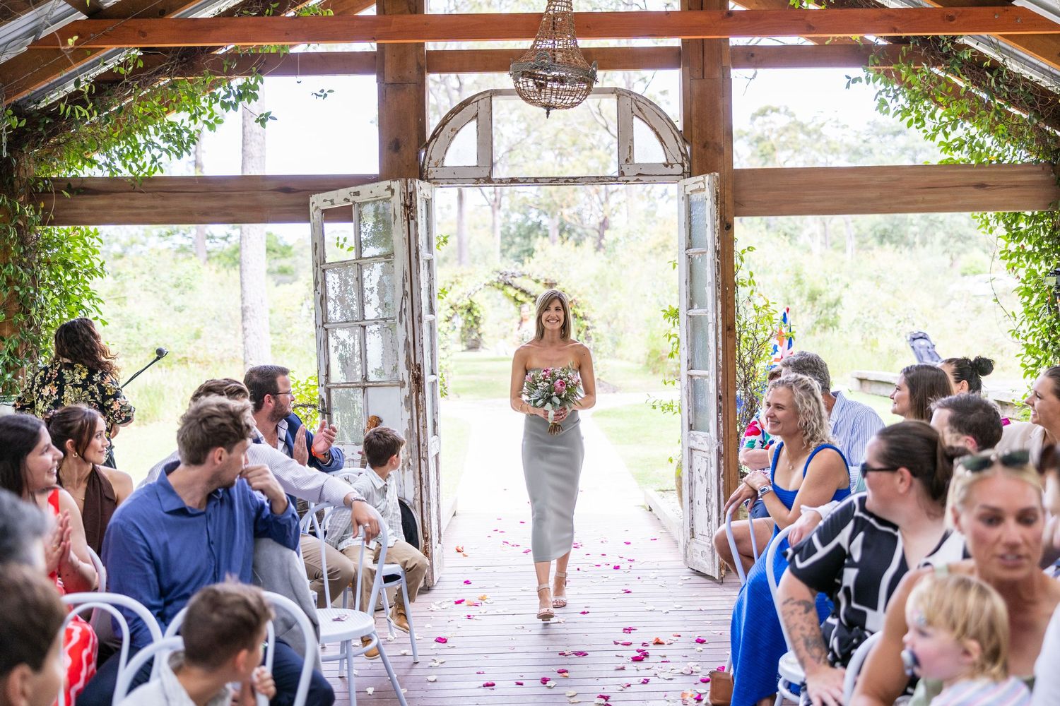 A bridesmaid in grey walks down a rose petal-lined aisle in a rustic chapel as wedding guests look on.
