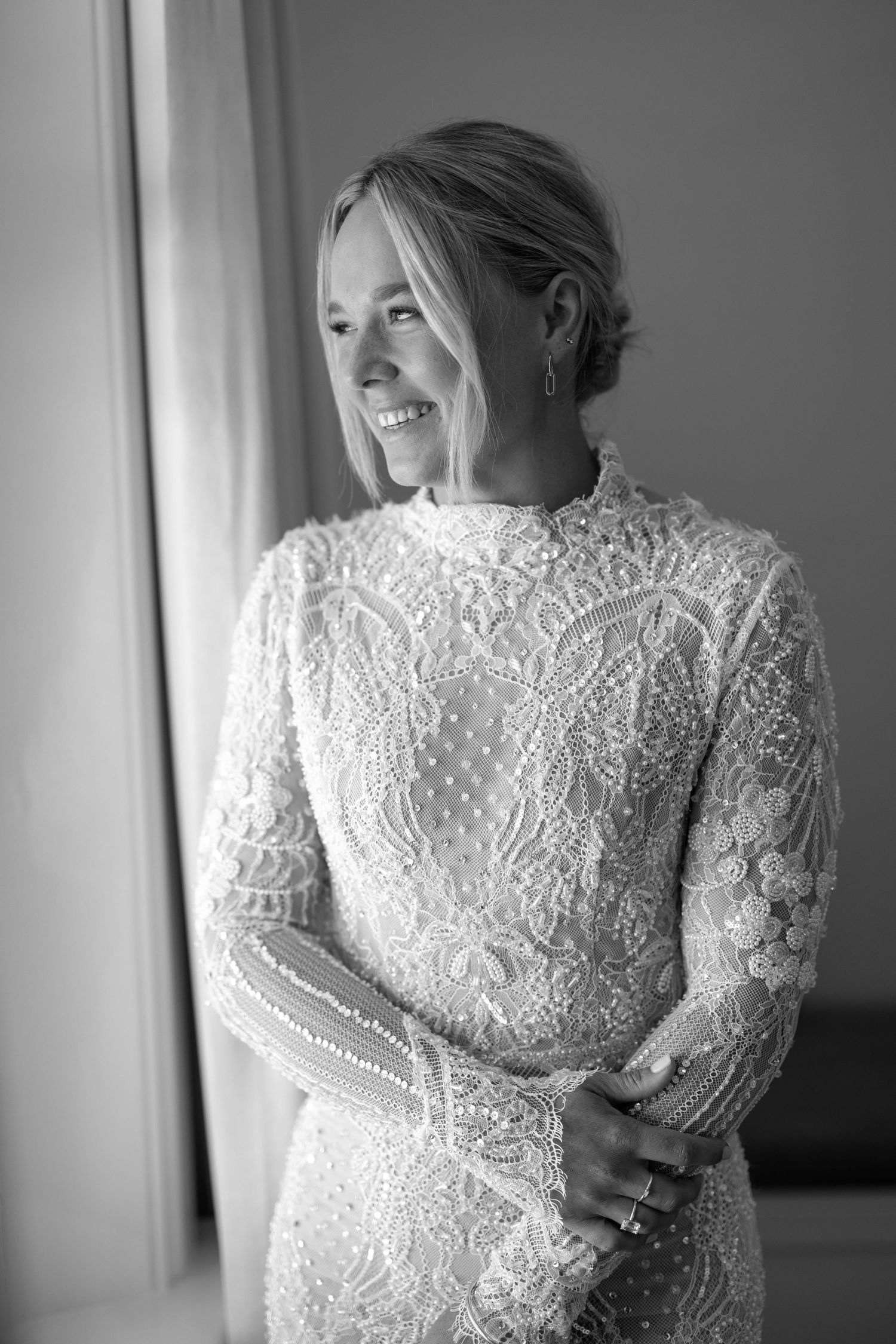 Black and white photo of a smiling bride in an elegant long-sleeve lace wedding dress, standing by a window.