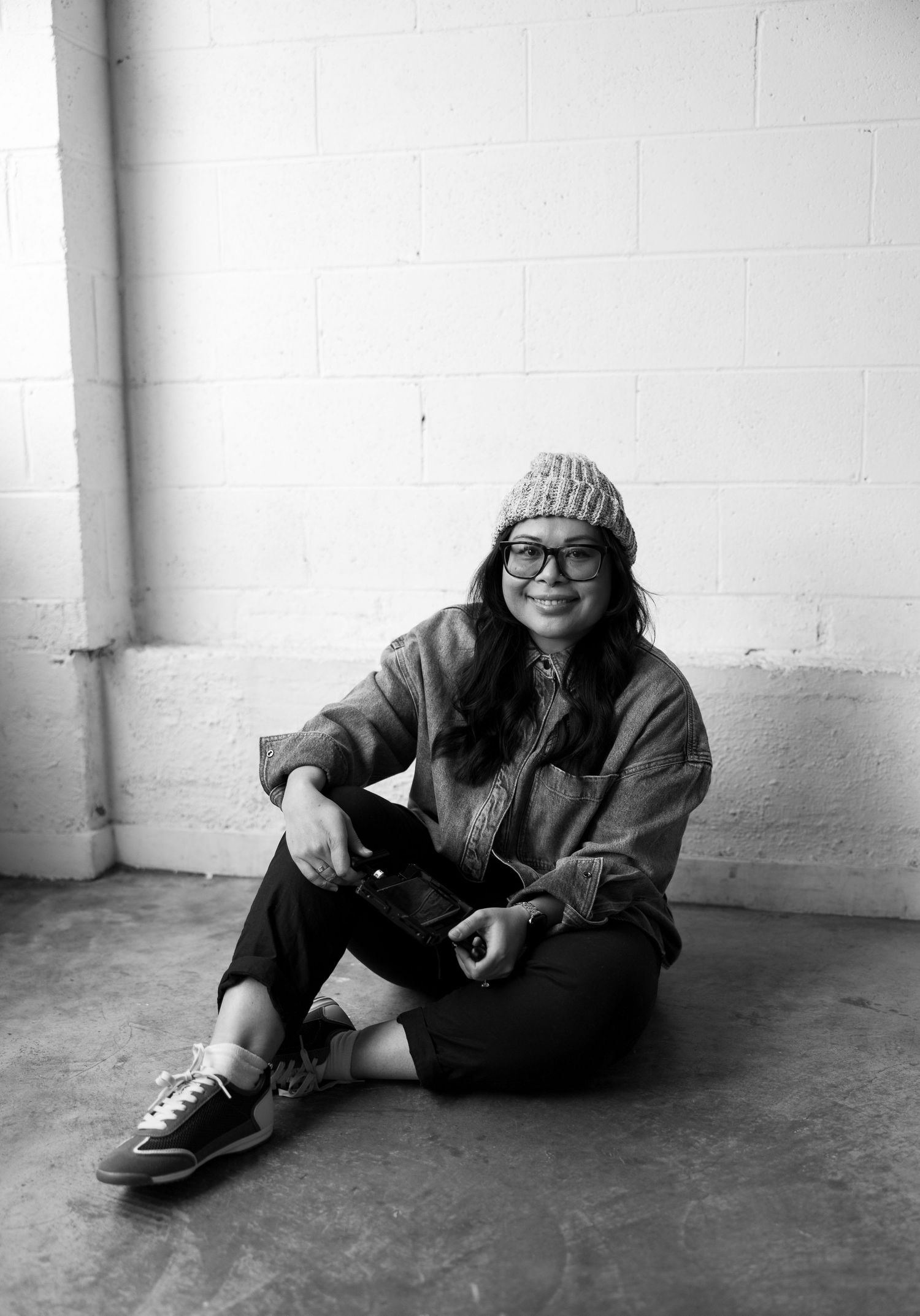 Black and white photo of a young woman sitting on floor, wearing glasses, beanie hat, and casual jacket, smiling.