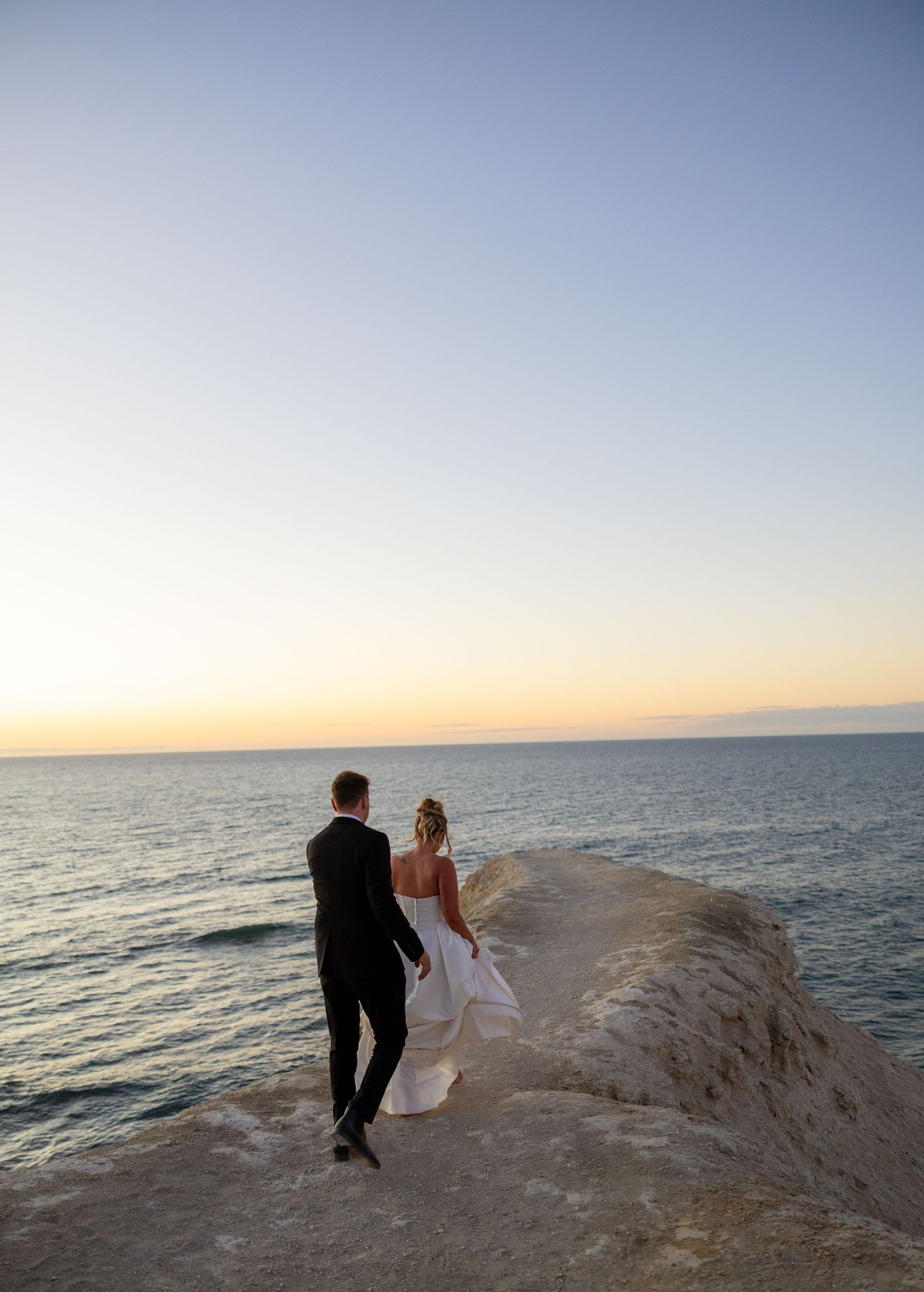 Bride and groom stand on coastal rock overlooking serene ocean horizon at golden hour sunset.