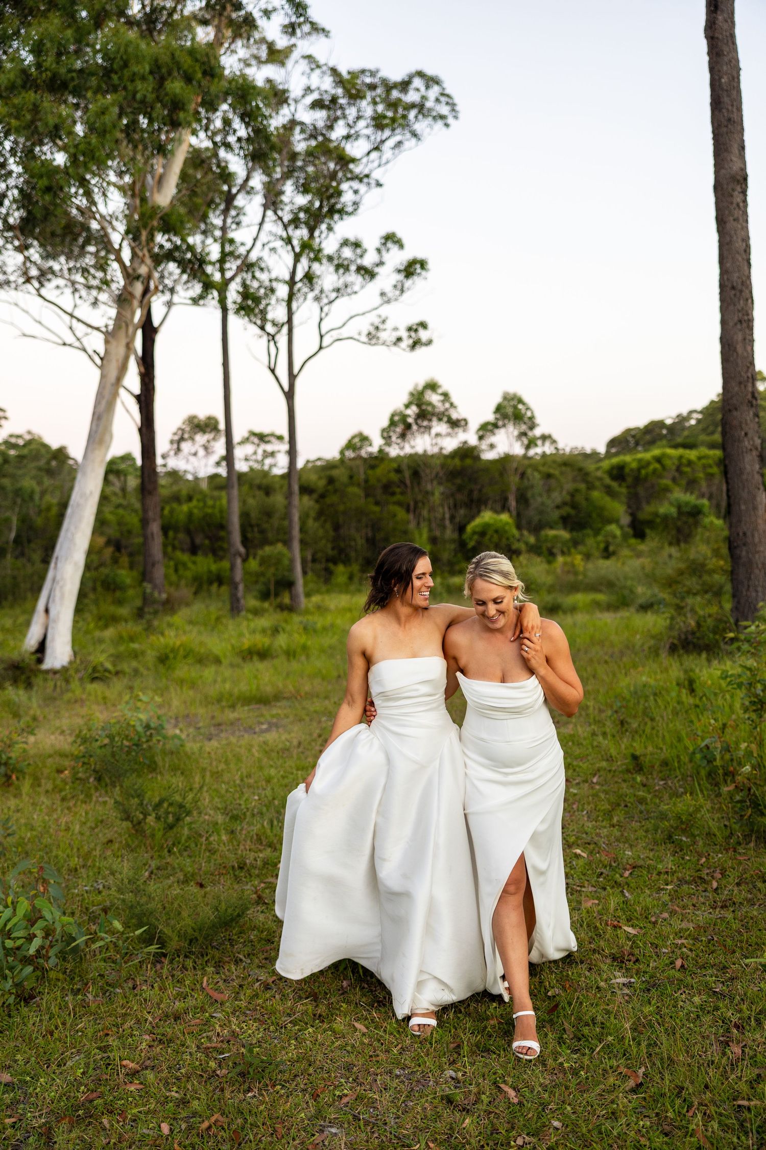 Two brides in white strapless gowns walk arm in arm through a lush green forest at sunset.