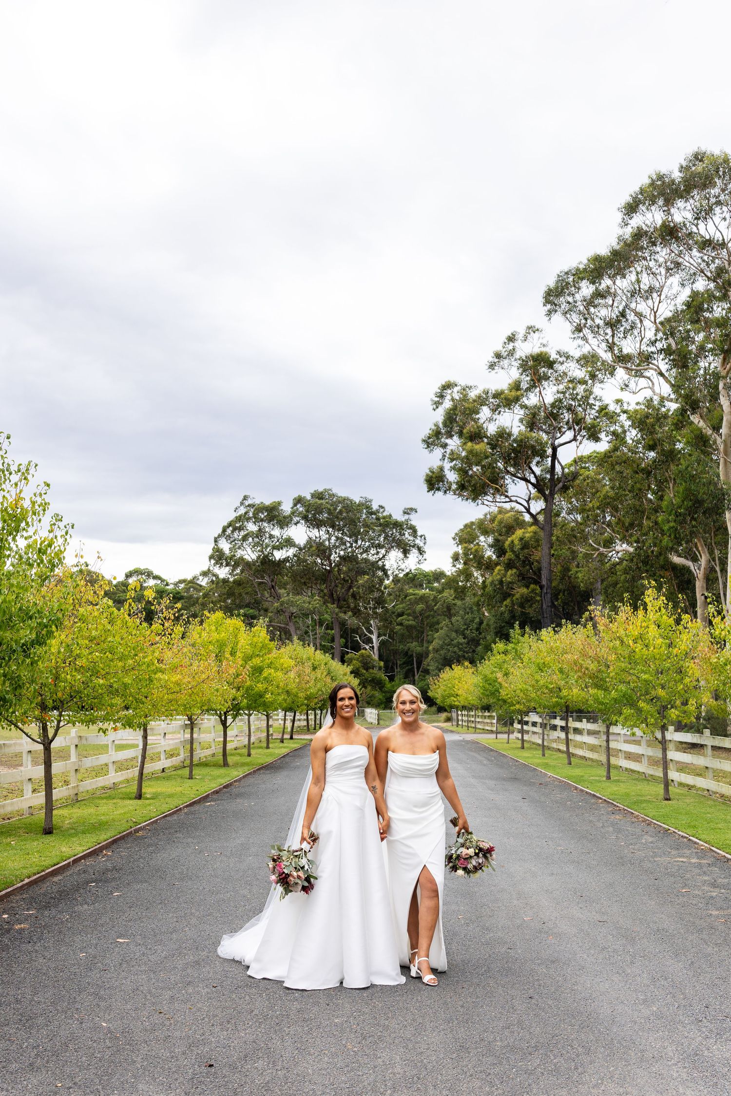 Two brides in white gowns walk hand-in-hand down a tree-lined driveway holding bouquets on their wedding day.