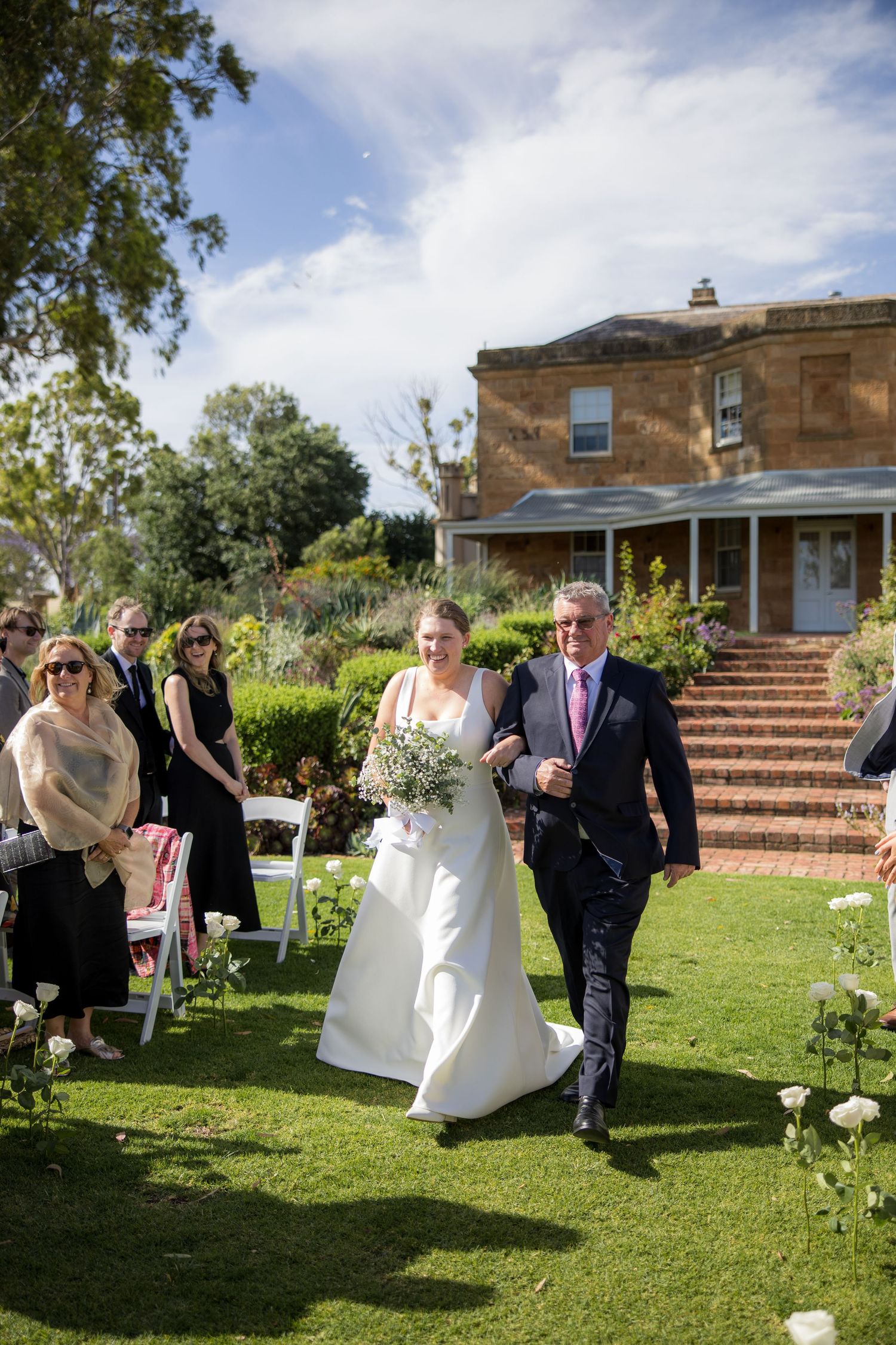 Bride walks down garden aisle with father at sunny outdoor wedding at historic estate.