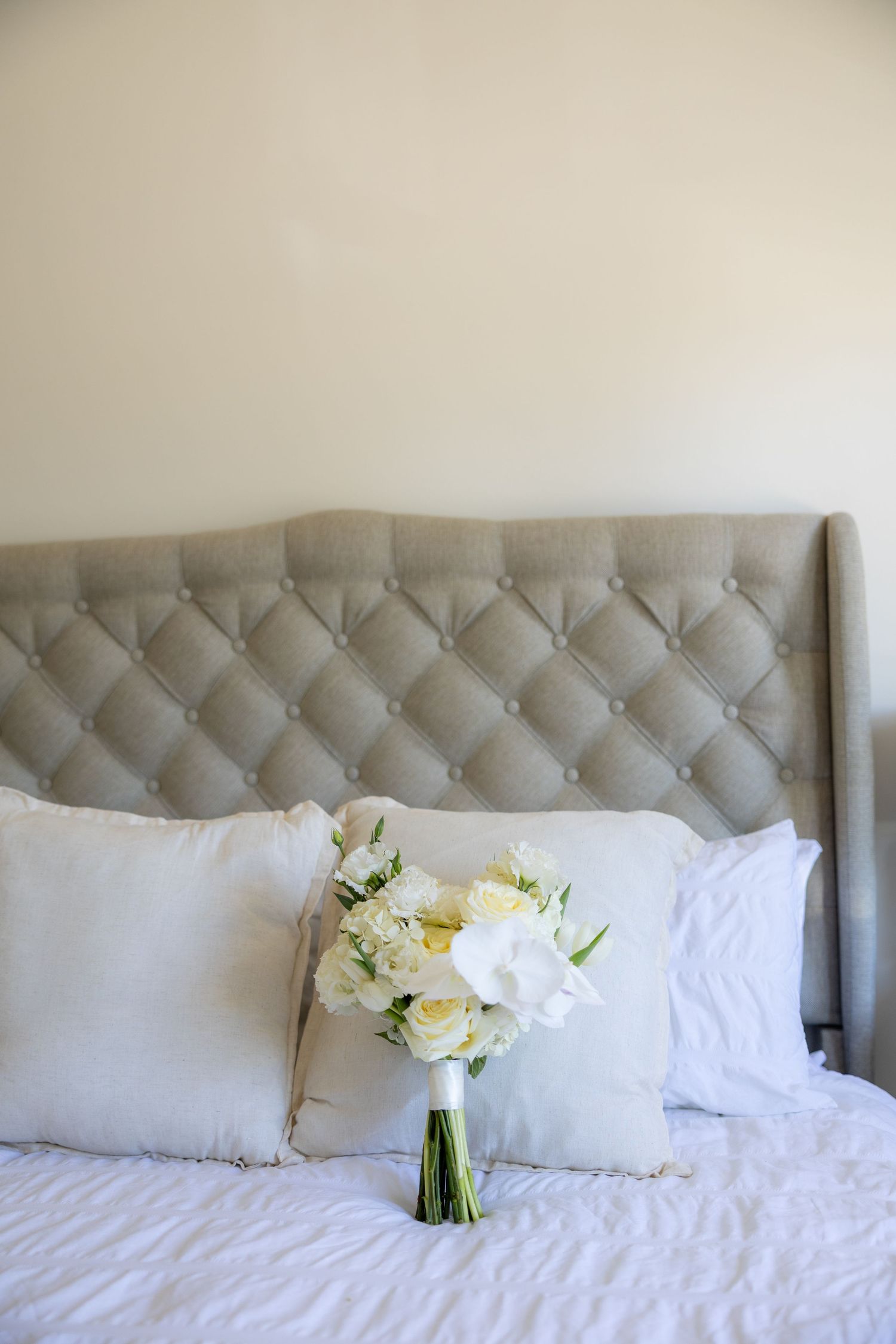White wedding bouquet of roses and peonies resting against pillows on a tufted headboard bed.