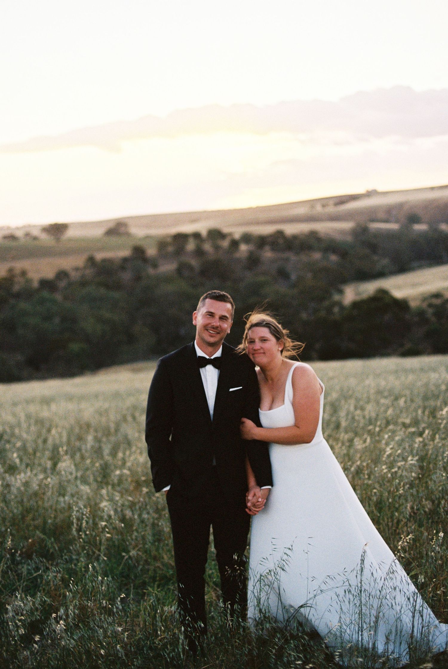 Bride and groom pose together in golden field at sunset, groom wearing black tuxedo with bow tie.