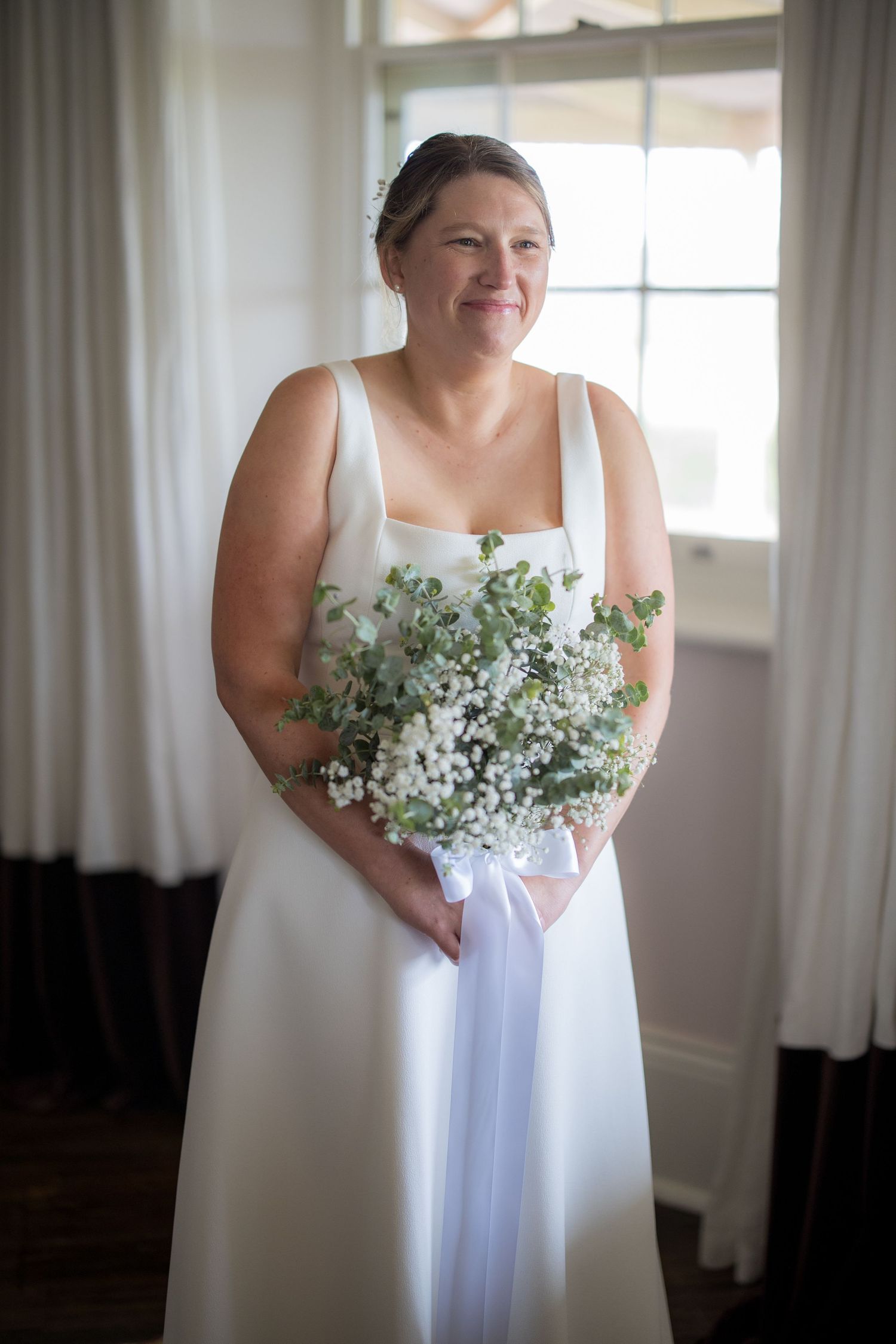 Bride in square-neck white gown holds a baby's breath bouquet near a sunlit window.