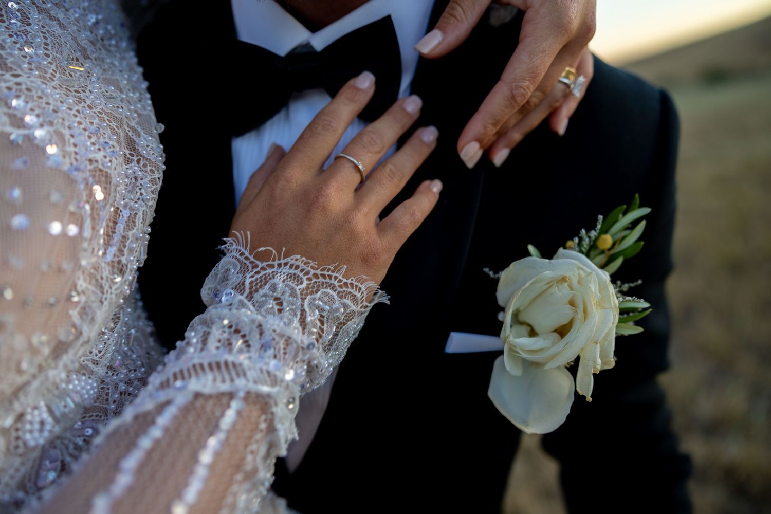 Detail shot of bride's beaded lace sleeve and diamond ring resting on groom's black tuxedo lapel.