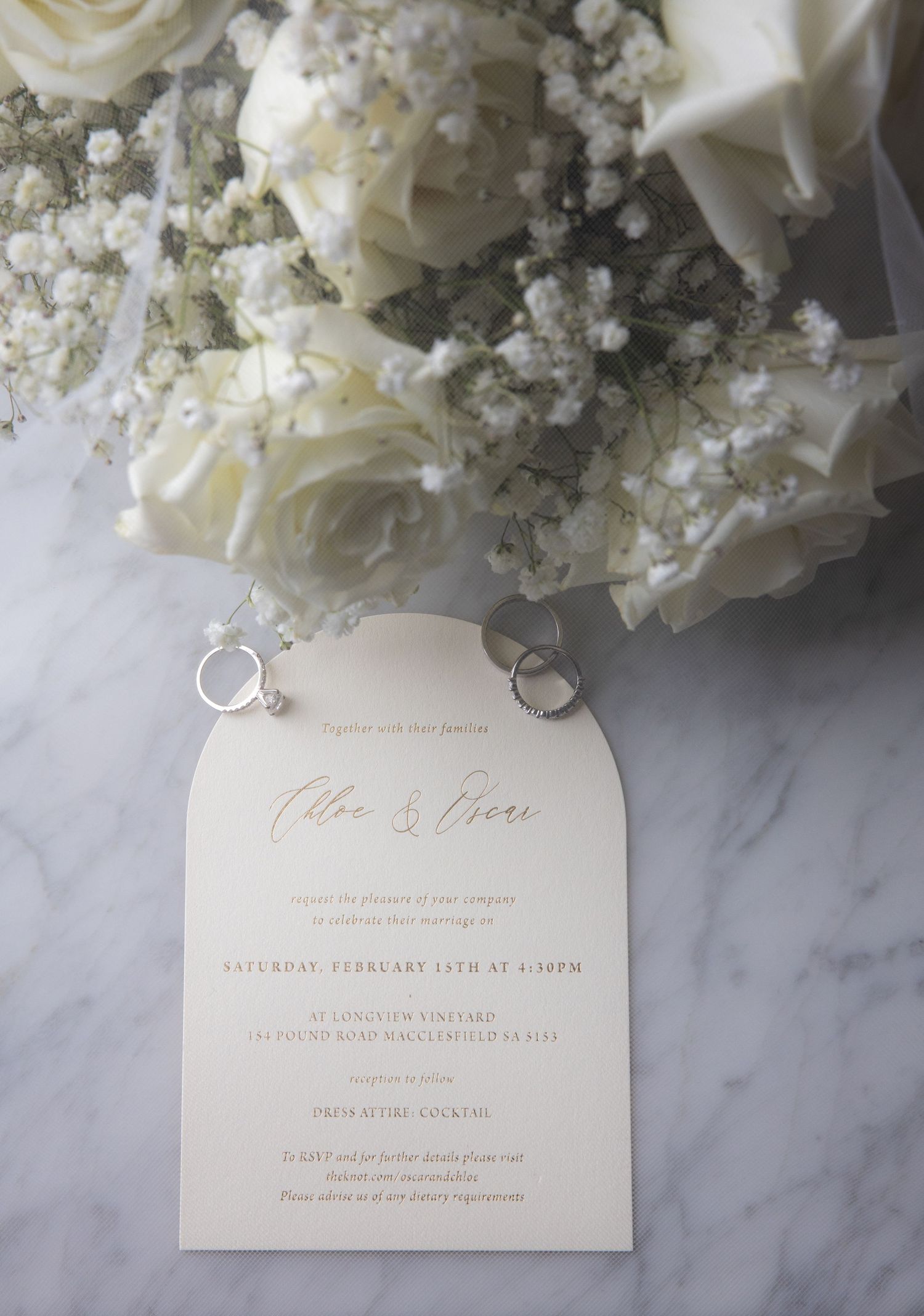Elegant wedding invitation with gold script beside white roses, baby's breath, and two silver rings on marble.