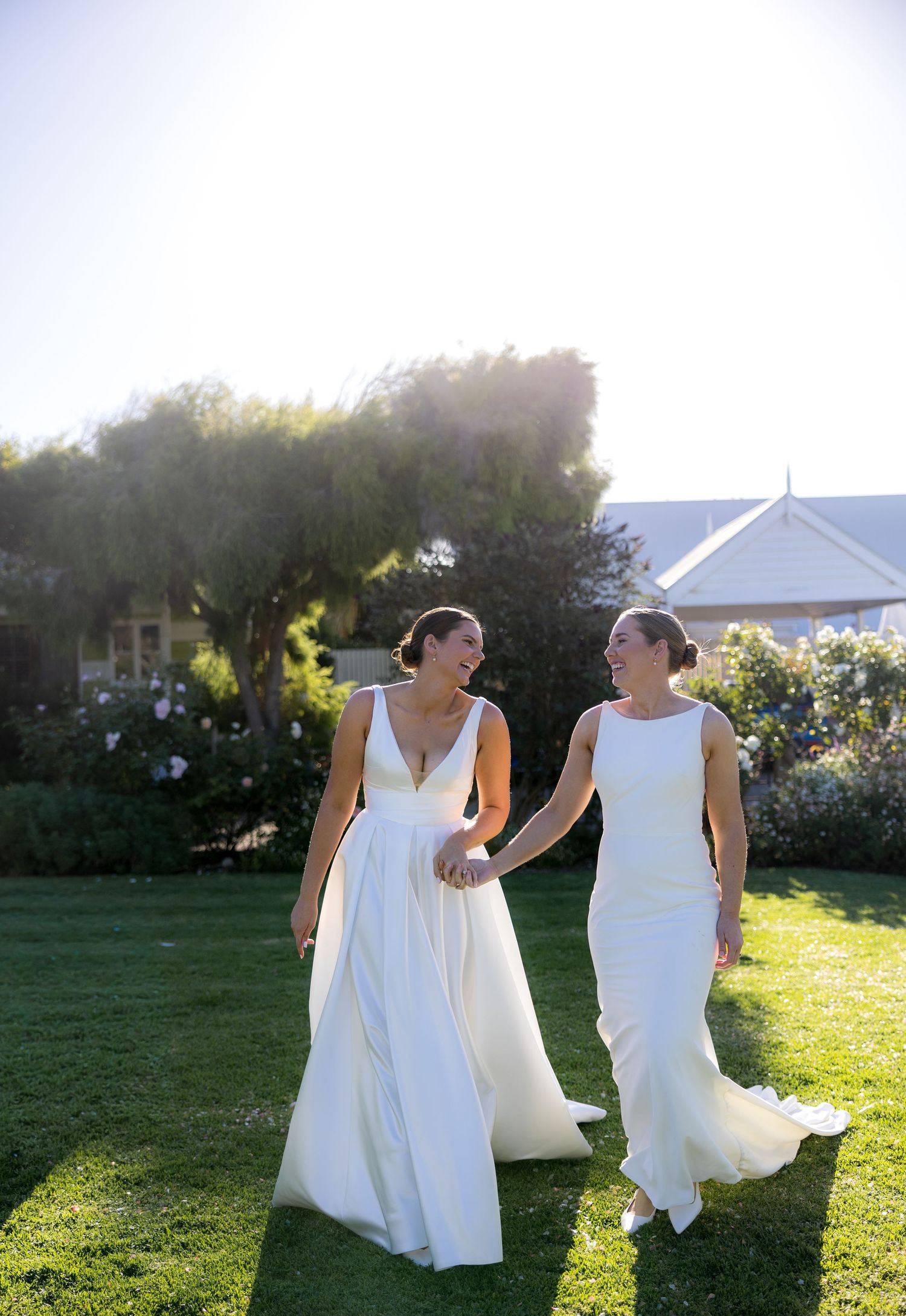 Two brides in elegant white gowns hold hands and walk across a sunlit green lawn after wedding.