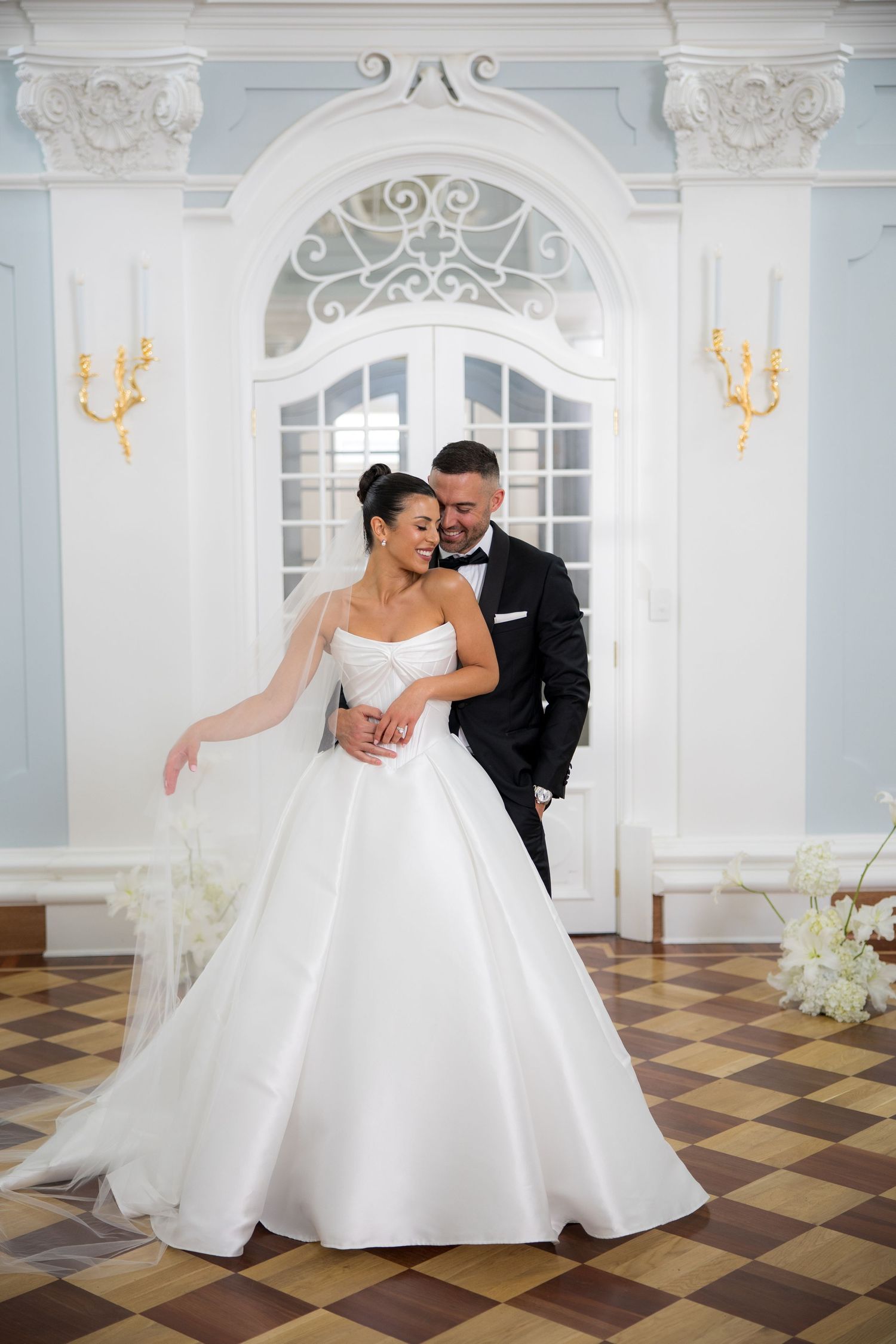 Bride in a strapless ballgown and groom in a black tuxedo dancing in an elegant blue and white ballroom.