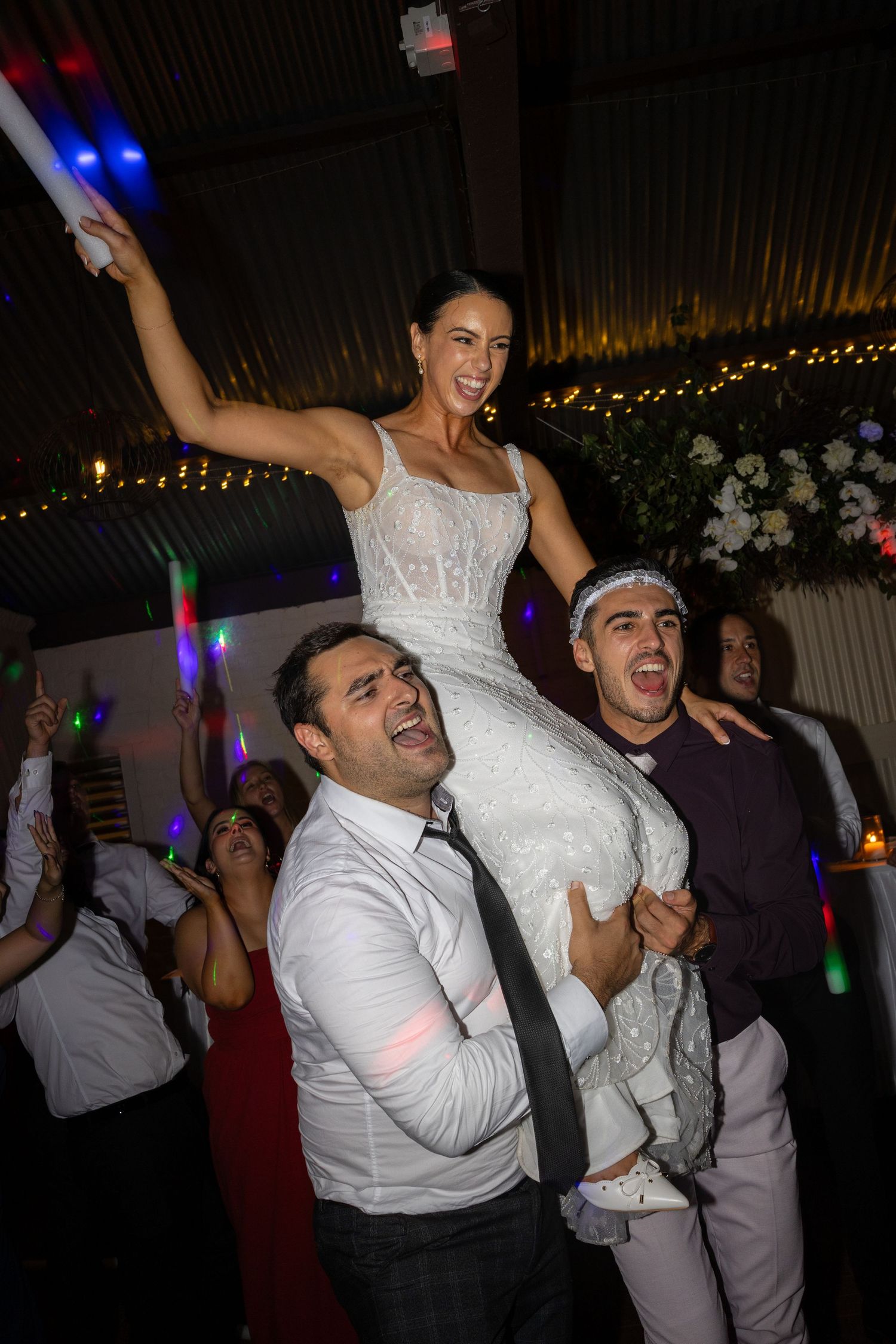 Bride in white lace dress lifted on shoulders by guests during lively wedding reception dance.