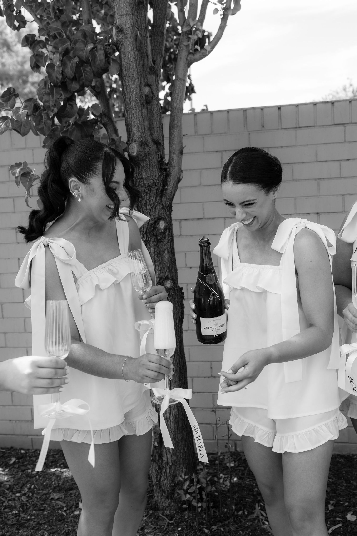 Two bridesmaids in white ruffled sets laughing while opening champagne bottle outdoors.
