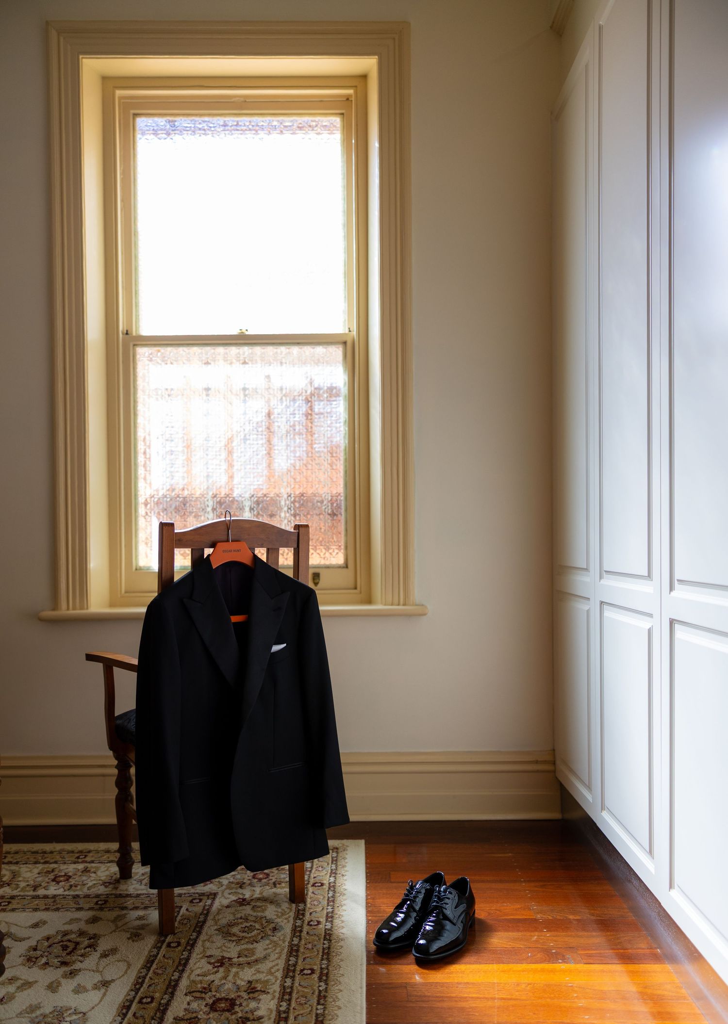 Black suit jacket on wooden chair with polished dress shoes by a sunlit window in a room.