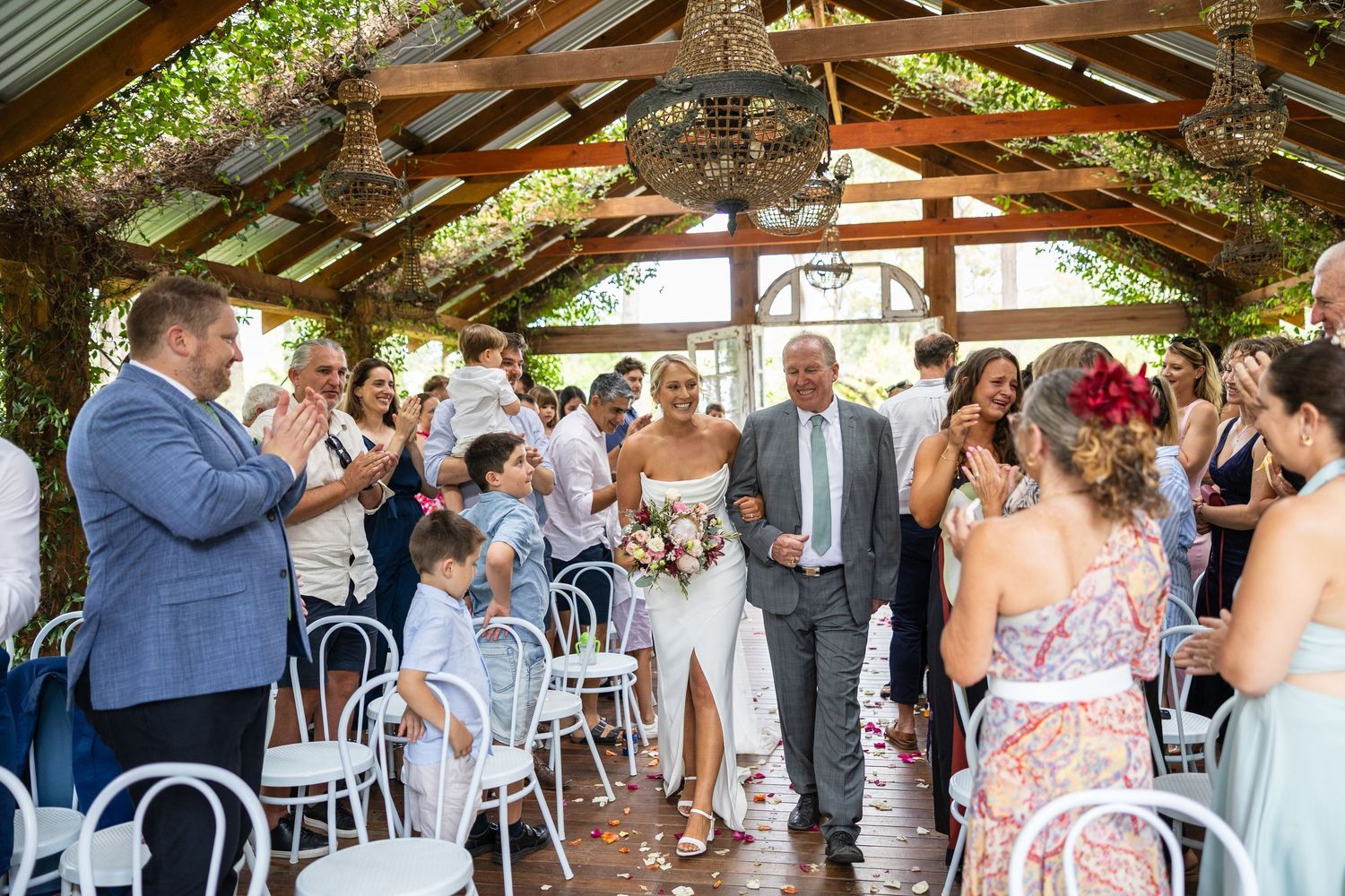 A bride walks down the aisle with an escort as smiling guests applaud in a rustic open-air wedding venue.