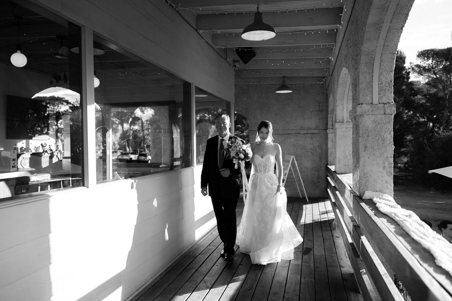 Black and white photo of bride and escort walking along sunlit wooden veranda of elegant wedding venue.