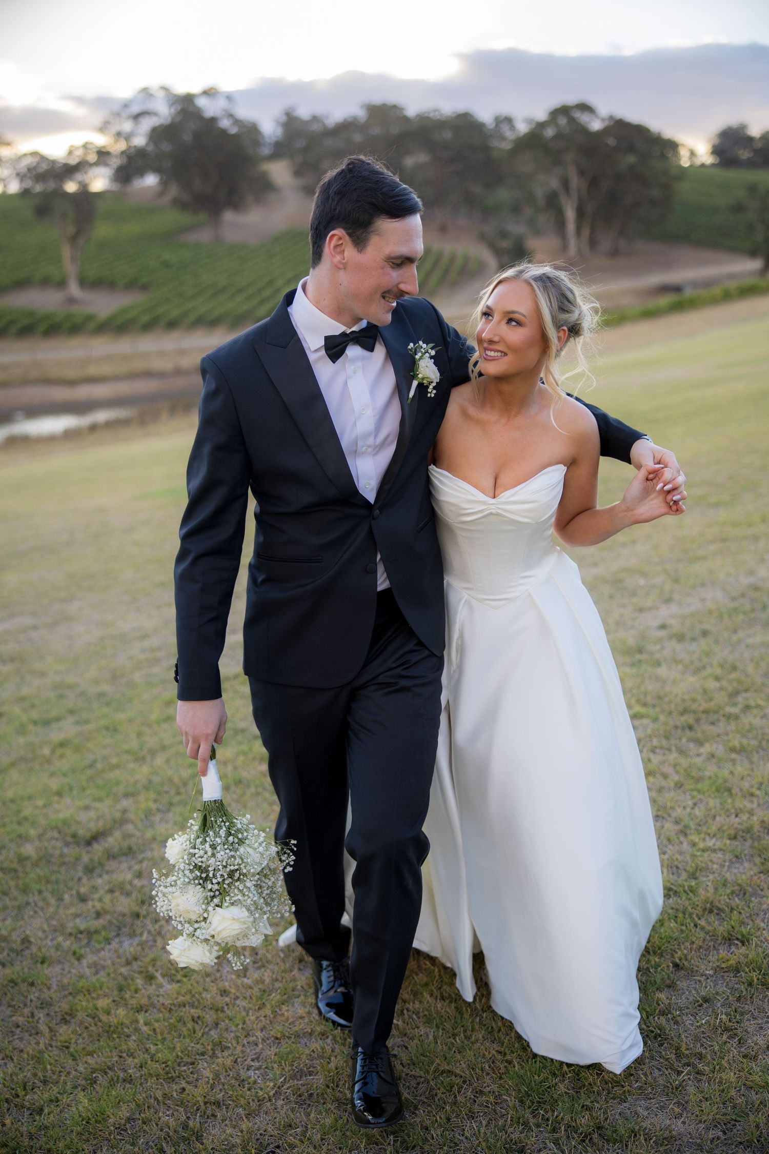 Bride in strapless gown smiles up at tall groom walking through vineyard at dusk.