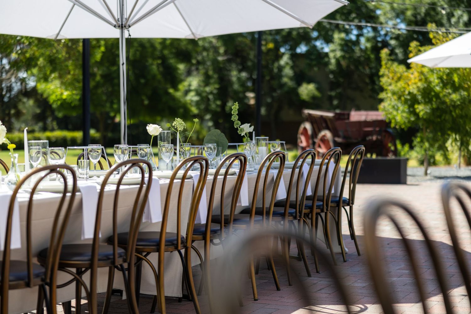 Elegant outdoor wedding reception table set with glassware and floral centerpieces under white umbrellas surrounded by lush greenery.