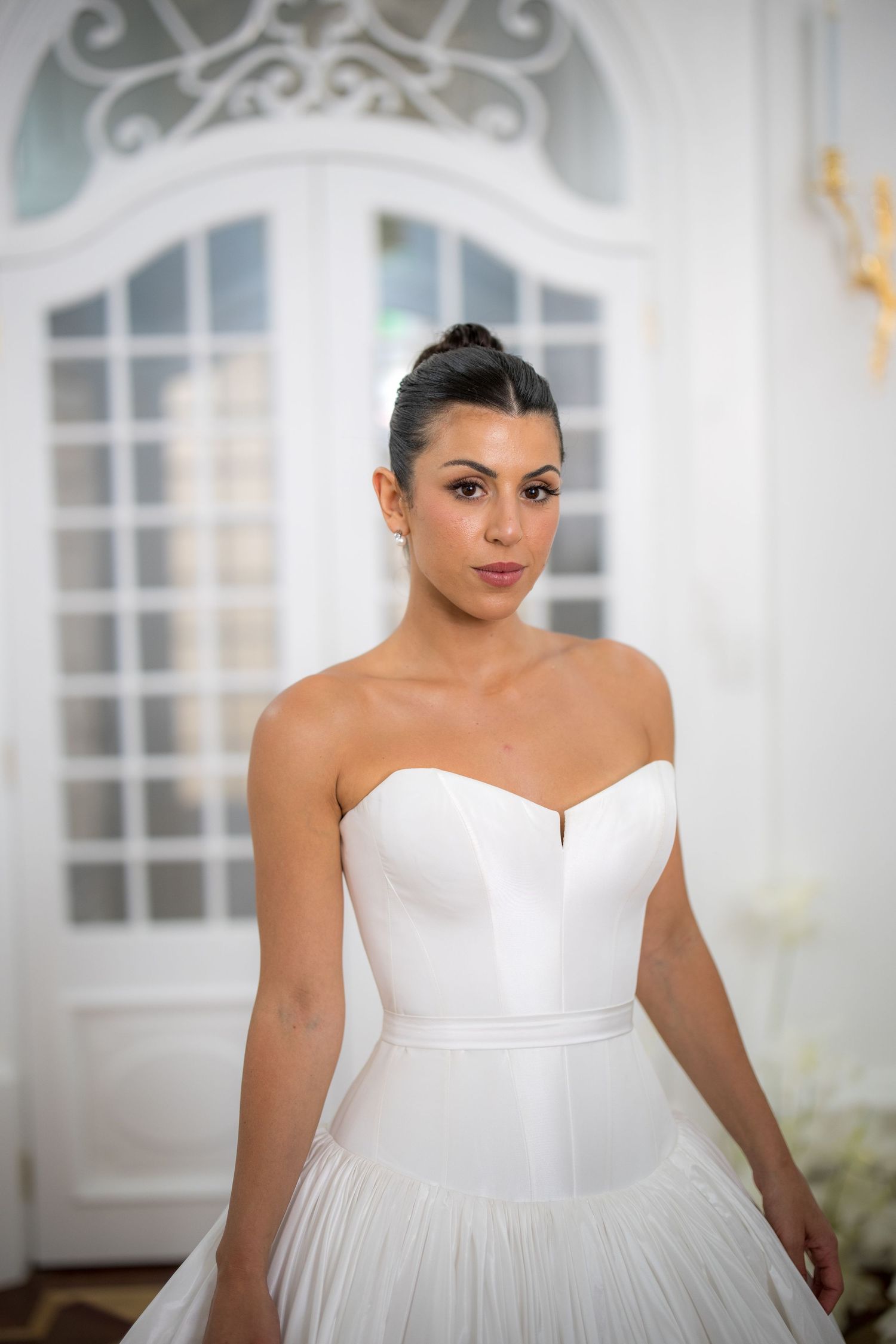 Elegant bride in strapless structured white ballgown posing before ornate white arched doors.
