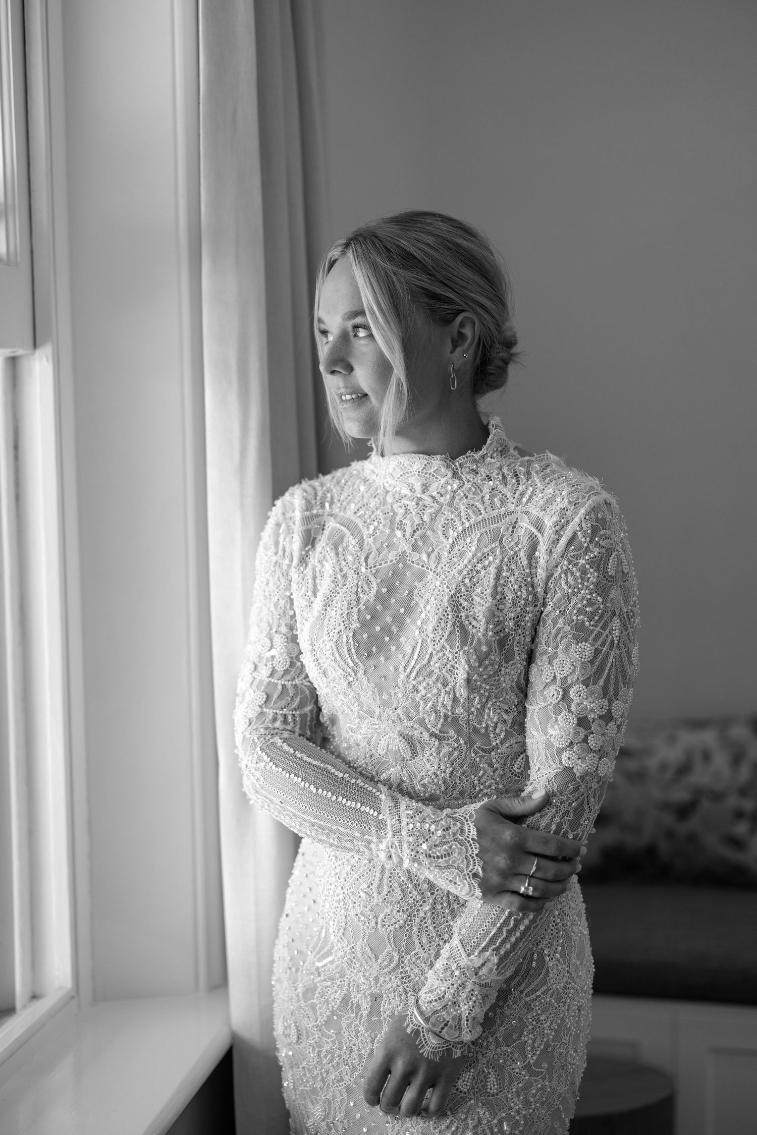 Smiling bride in long-sleeve lace wedding gown stands by window in elegant black and white portrait photo.
