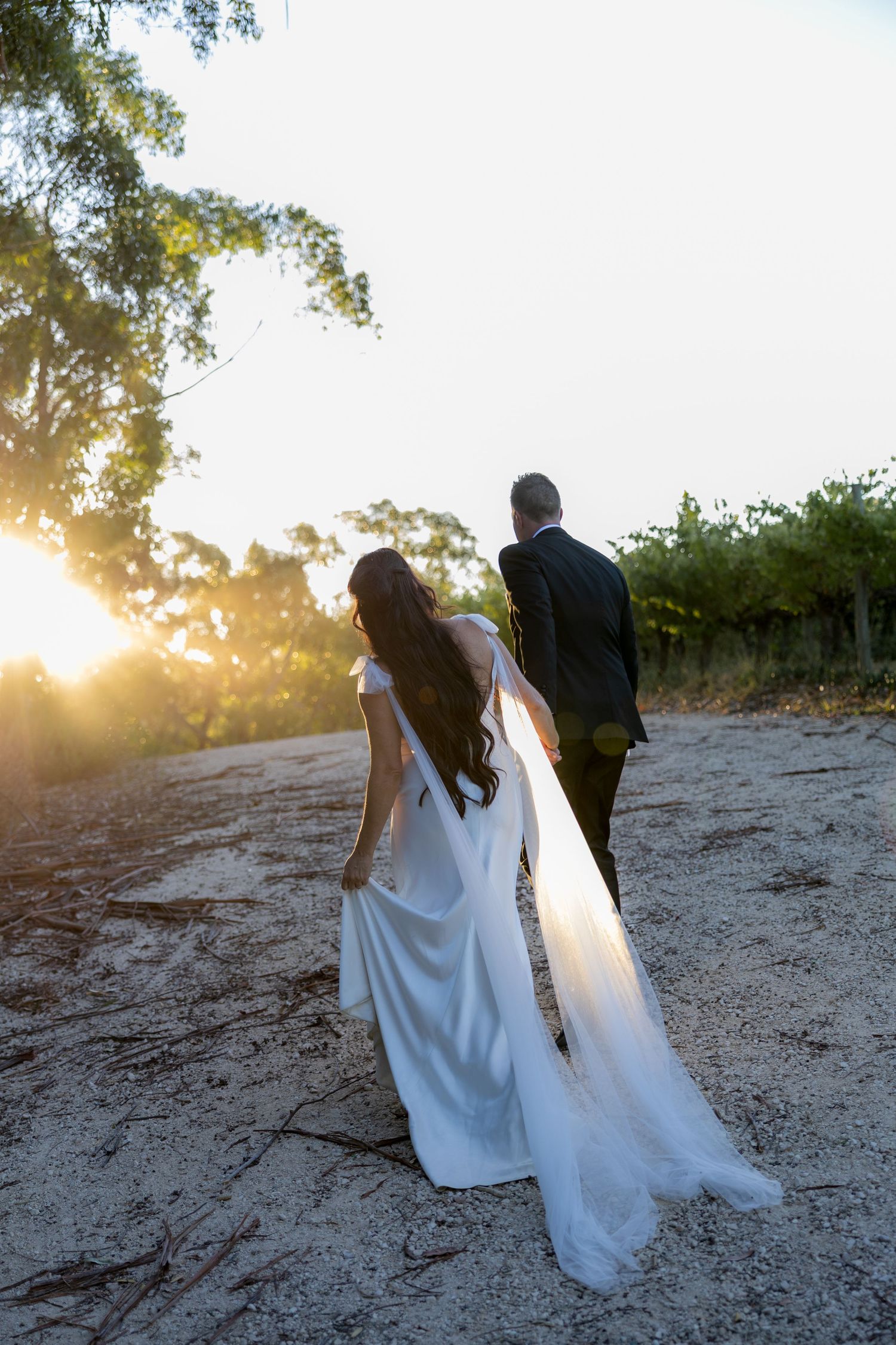Bride and groom walking toward sunset on sandy path surrounded by lush green trees.