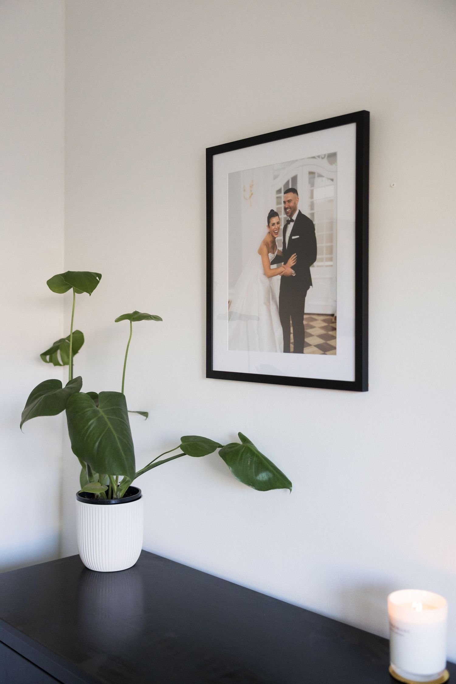 Black framed wedding photo displayed on white wall beside monstera plant on dark dresser.