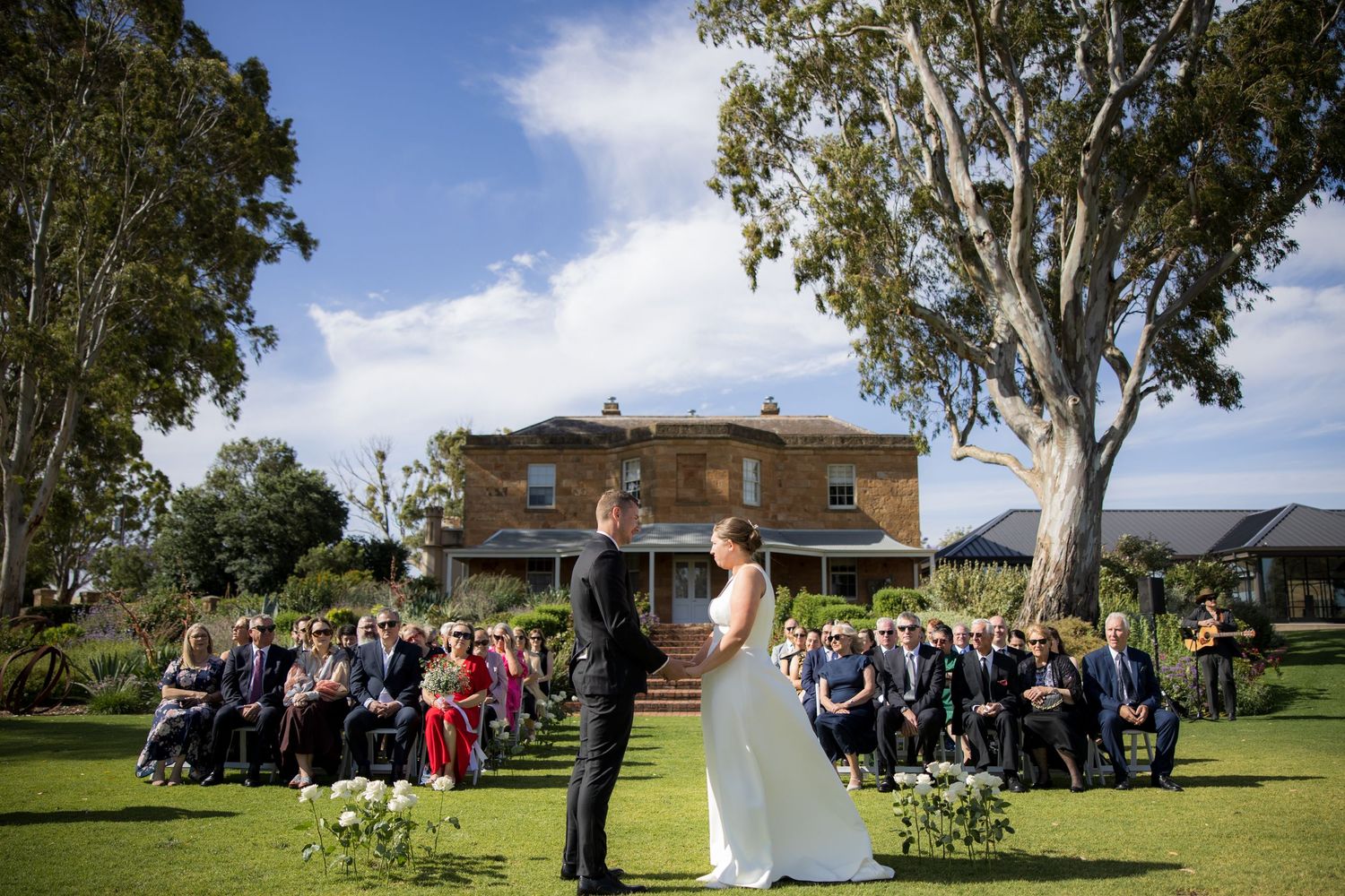 Bride and groom exchange vows at outdoor ceremony with historic stone estate backdrop.