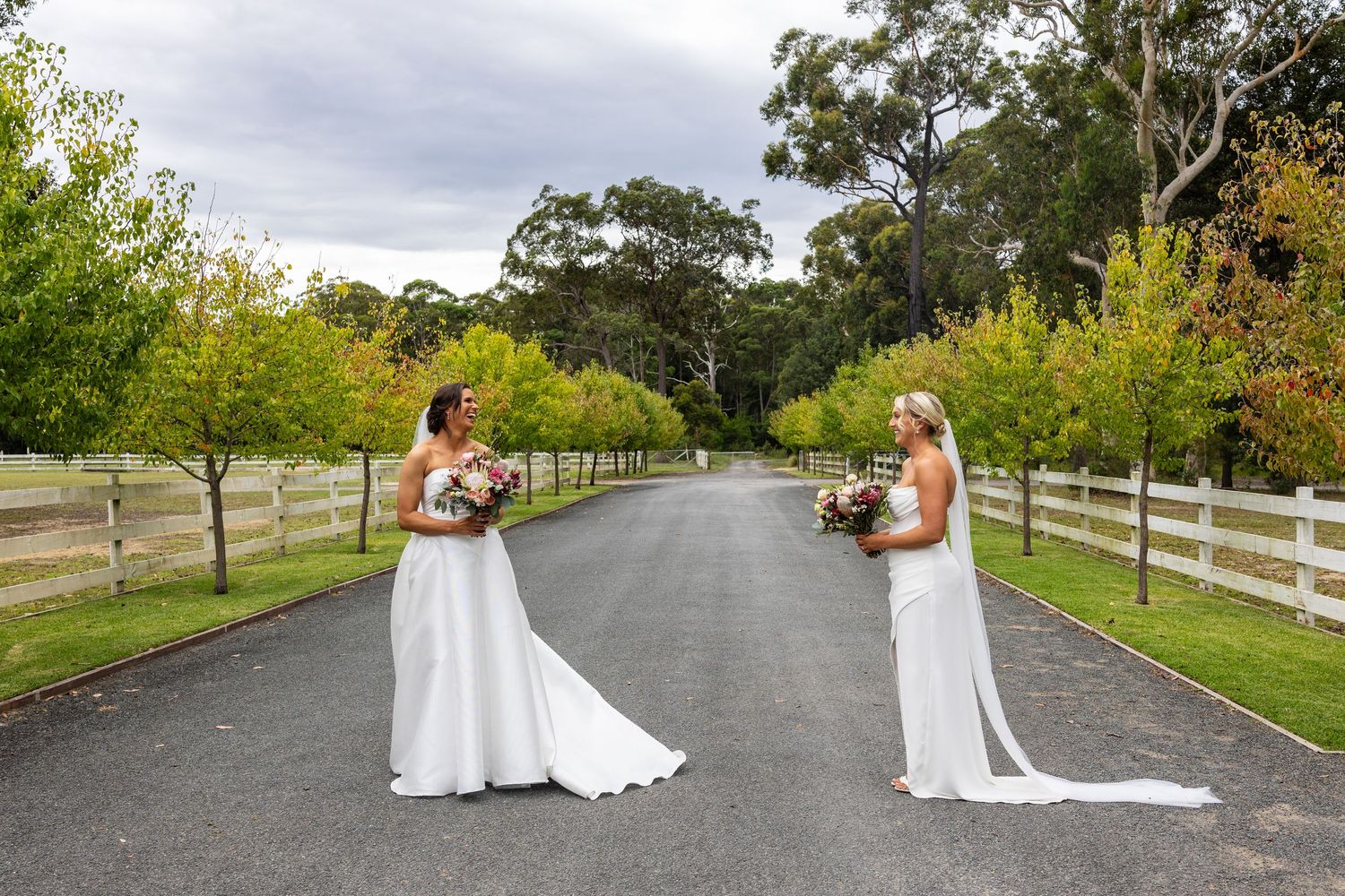 Two brides in white gowns face each other holding bouquets on a scenic tree-lined driveway.