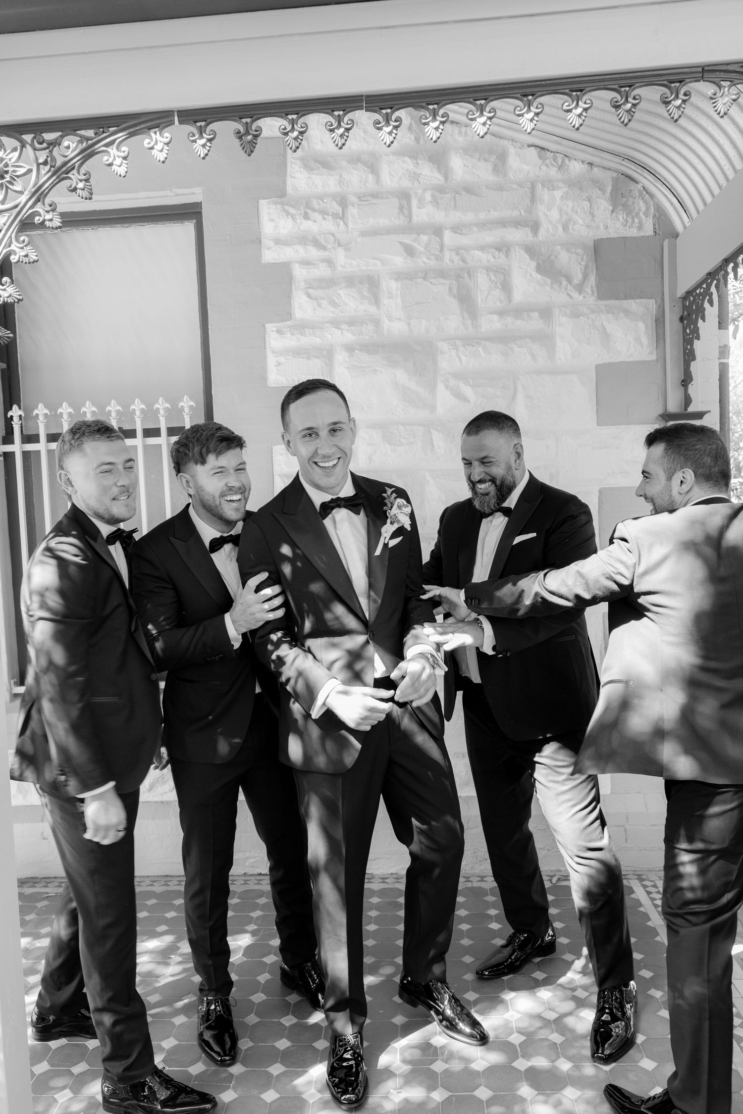Groom laughing with groomsmen in black tuxedos outside a heritage building in black and white photo.
