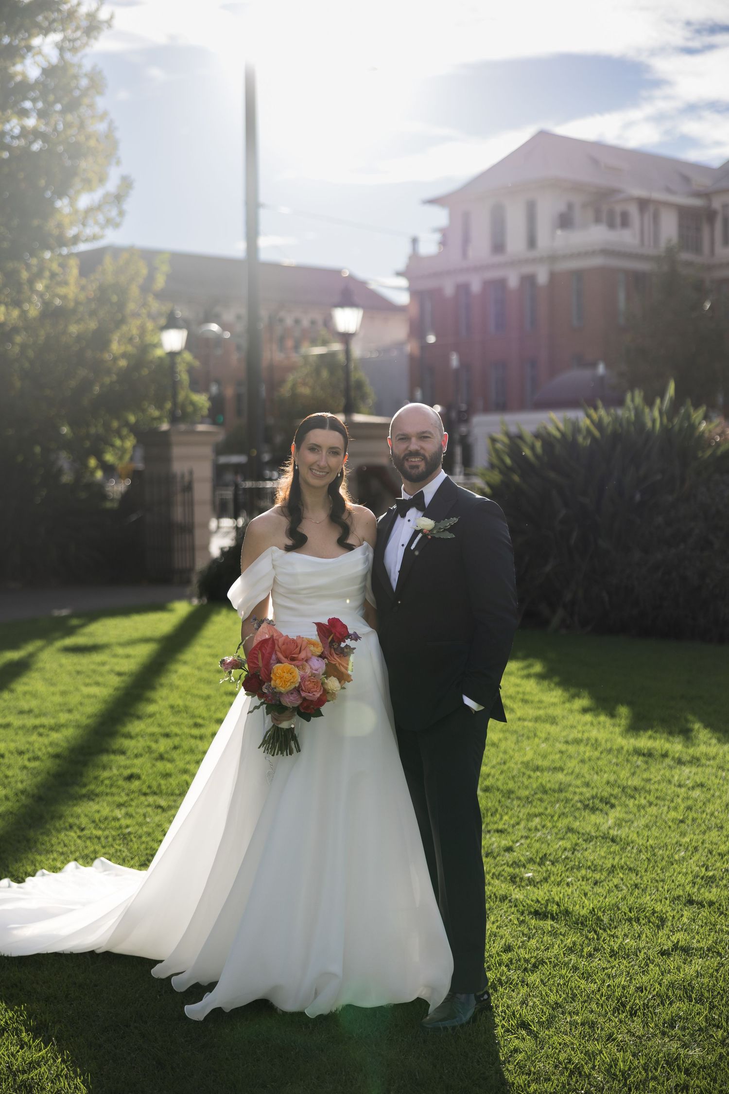 Bride in off-shoulder gown holding colorful bouquet stands with tuxedo-clad groom on green lawn outside historic building.