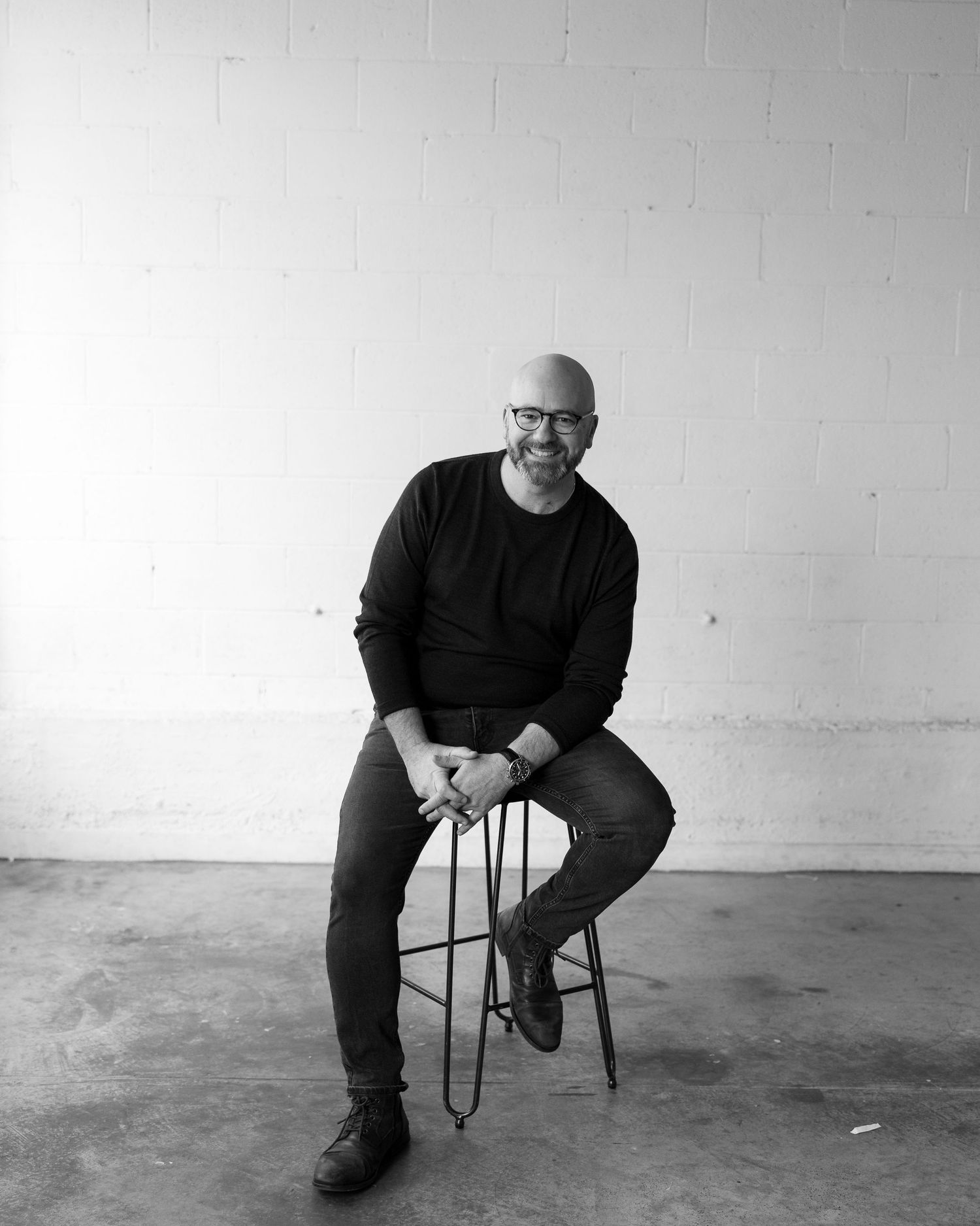 Bald man with glasses smiling, seated on a stool in a bright studio, wearing a black sweater and jeans.