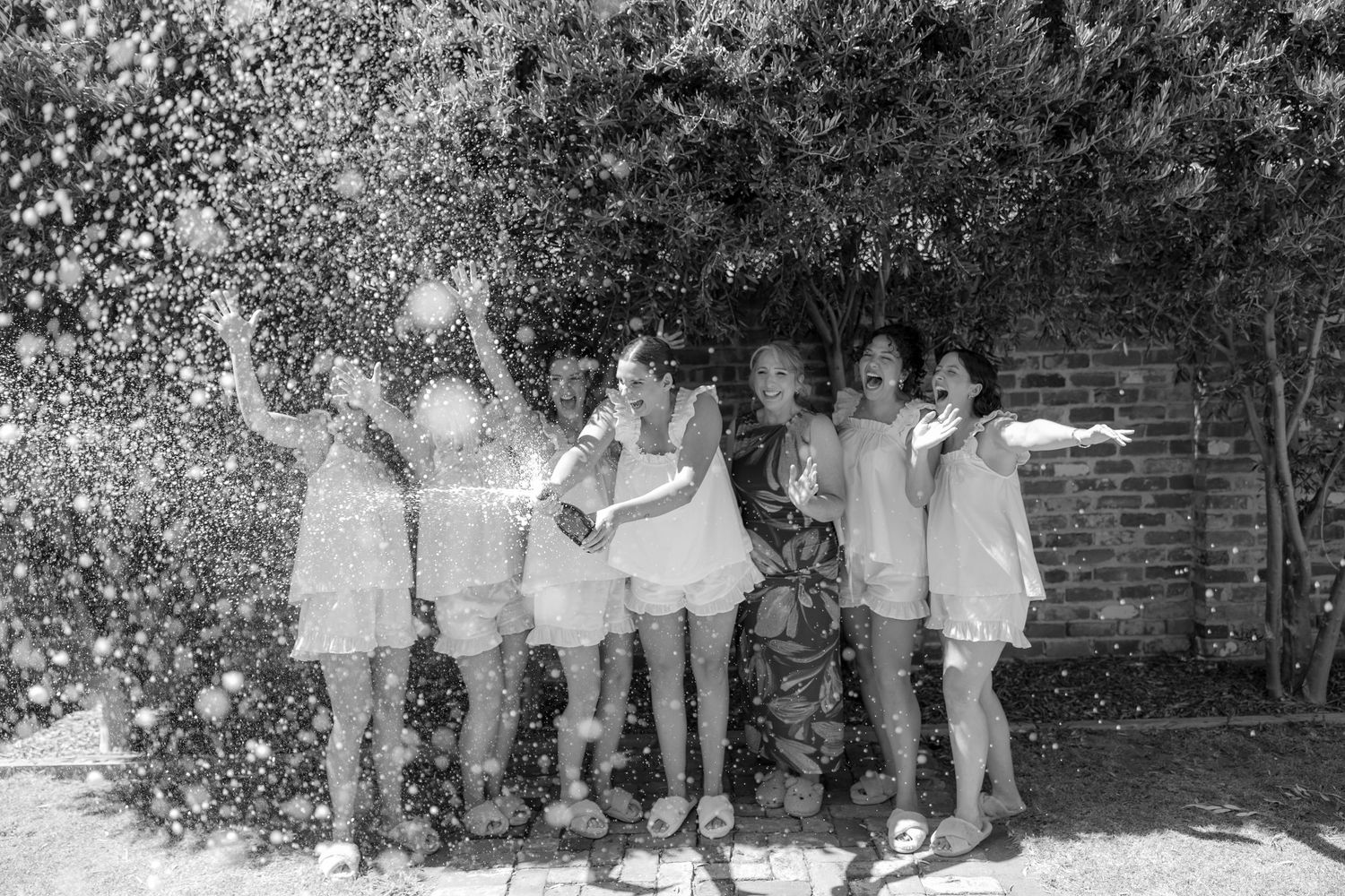 Black and white photo of bridal party celebrating with confetti explosion in sunny garden.