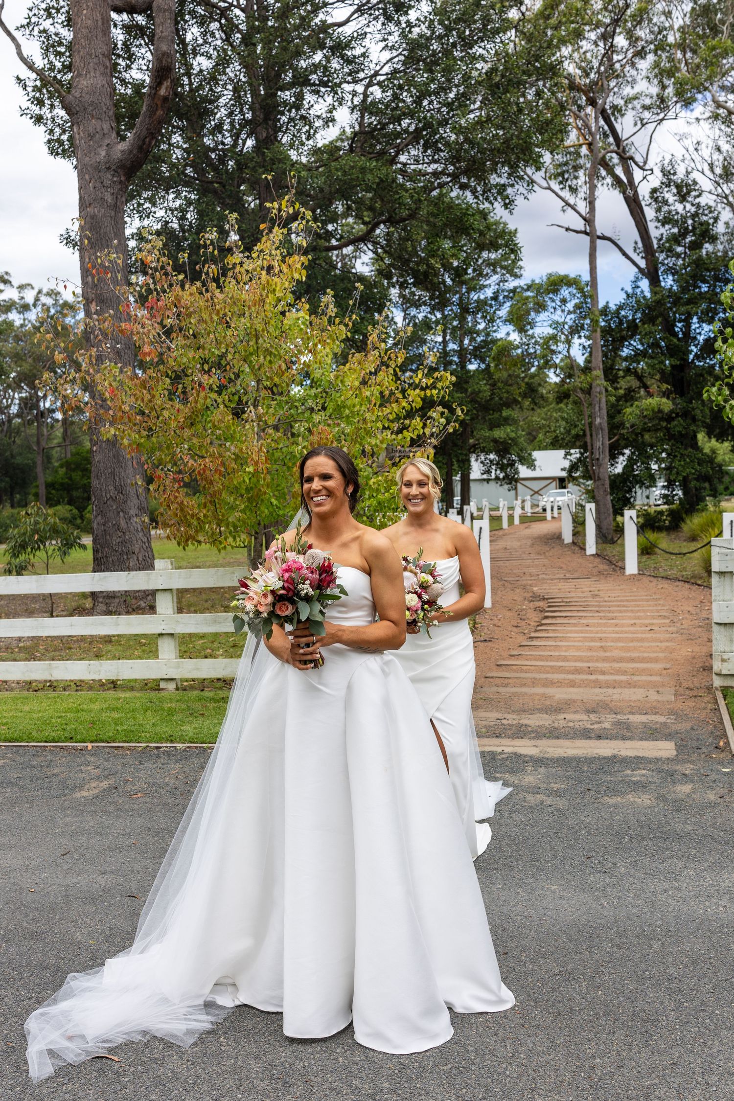 Two brides in elegant white gowns hold colorful bouquets while walking along a tree-lined driveway.