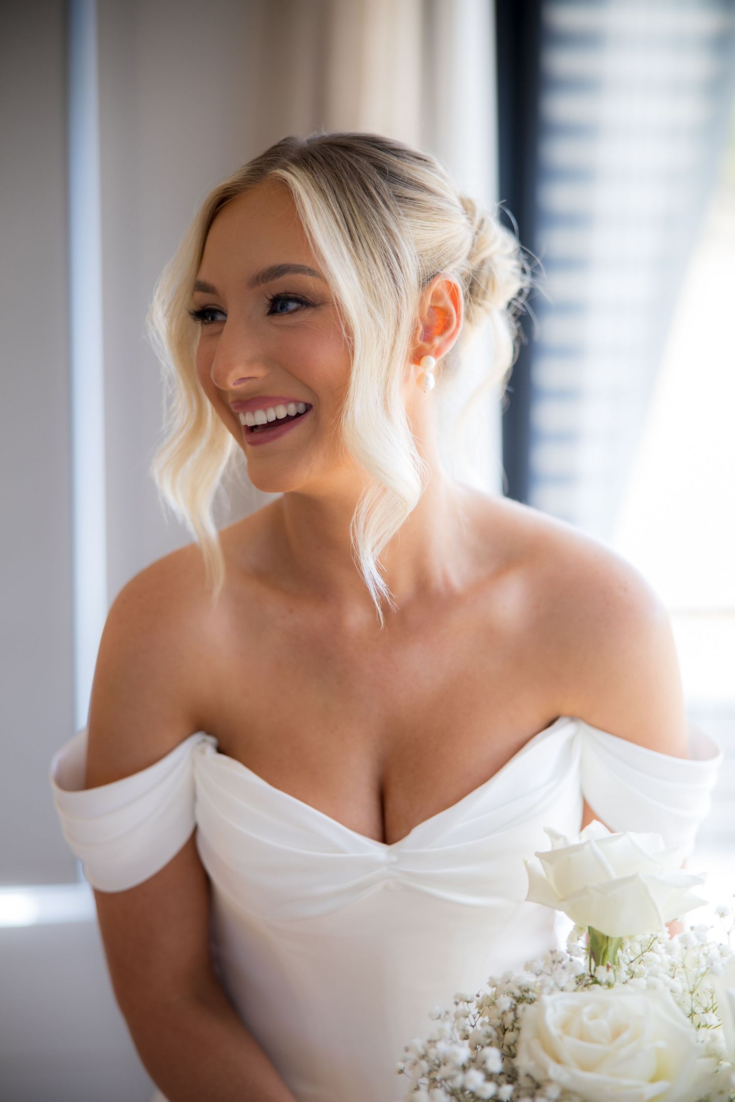 Radiant bride smiling joyfully in an off-shoulder white wedding dress holding white roses.