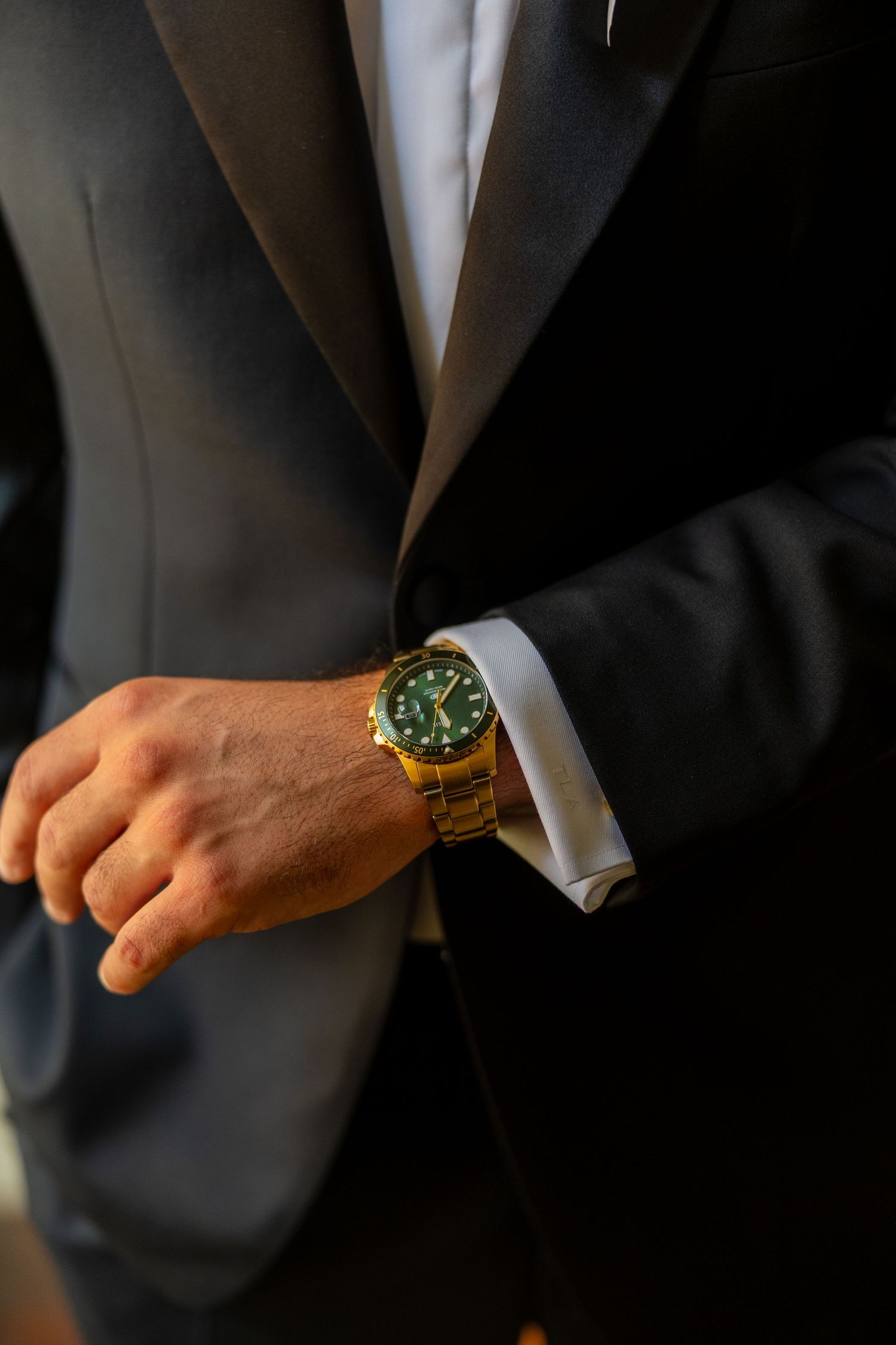 Groom adjusting his gold watch with green dial while wearing a black tuxedo and white dress shirt.