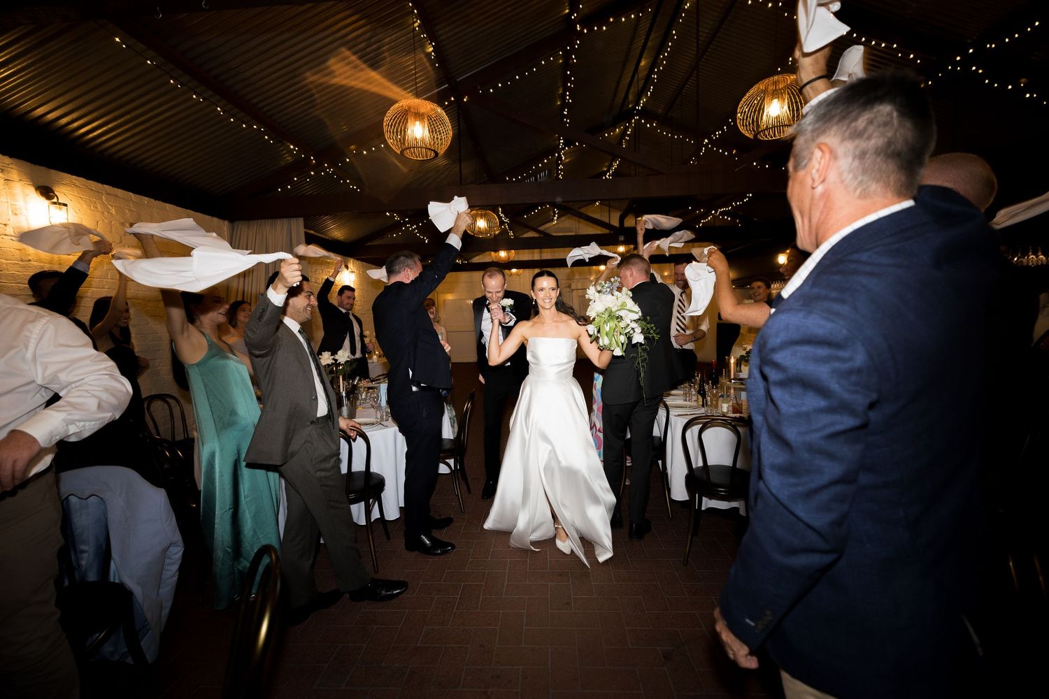 Bride in a white gown holding a bouquet walks through a wedding reception as smiling guests wave white napkins in celebration.