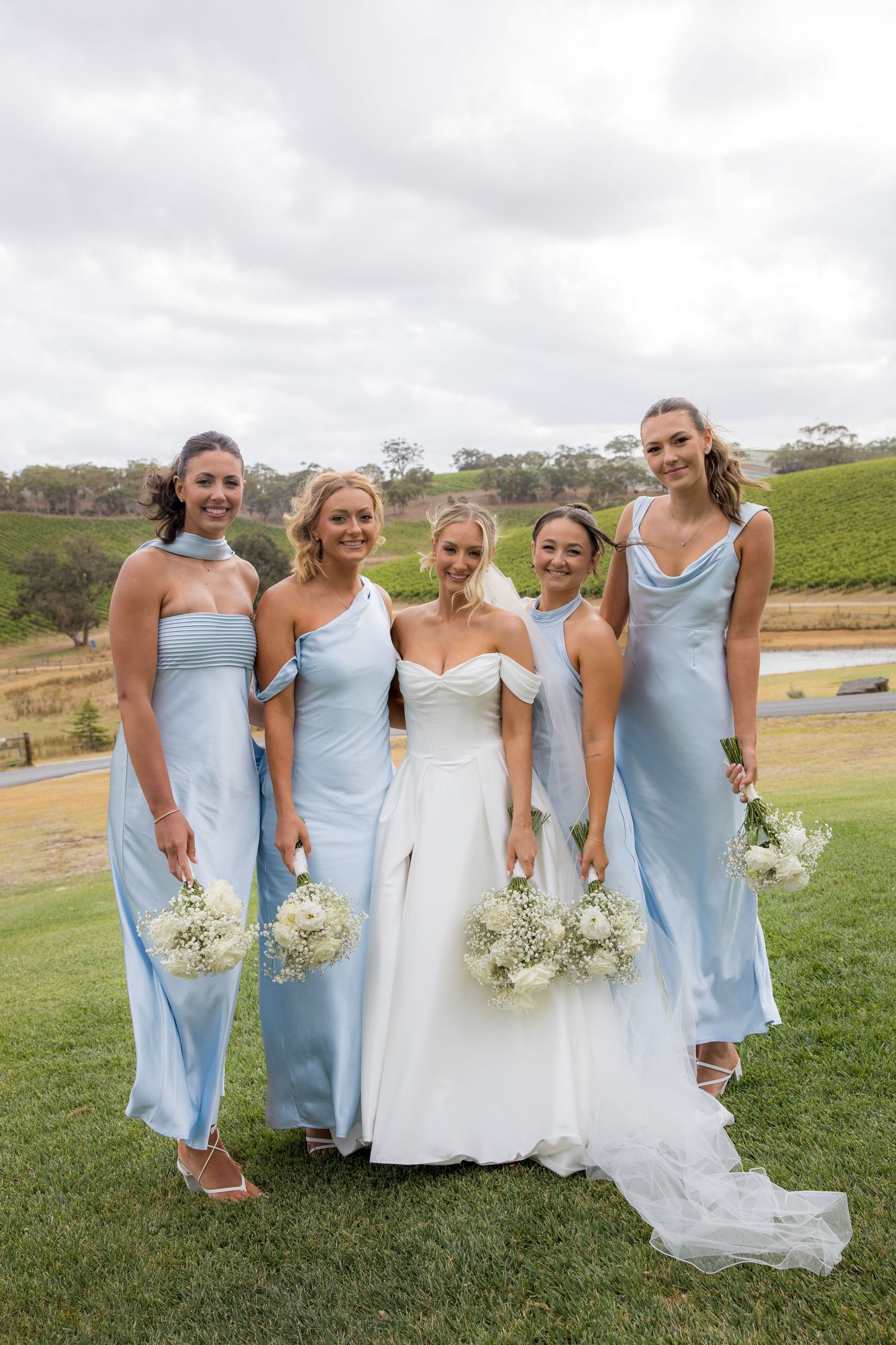 Bride in white gown poses with four bridesmaids in light blue dresses holding white bouquets.