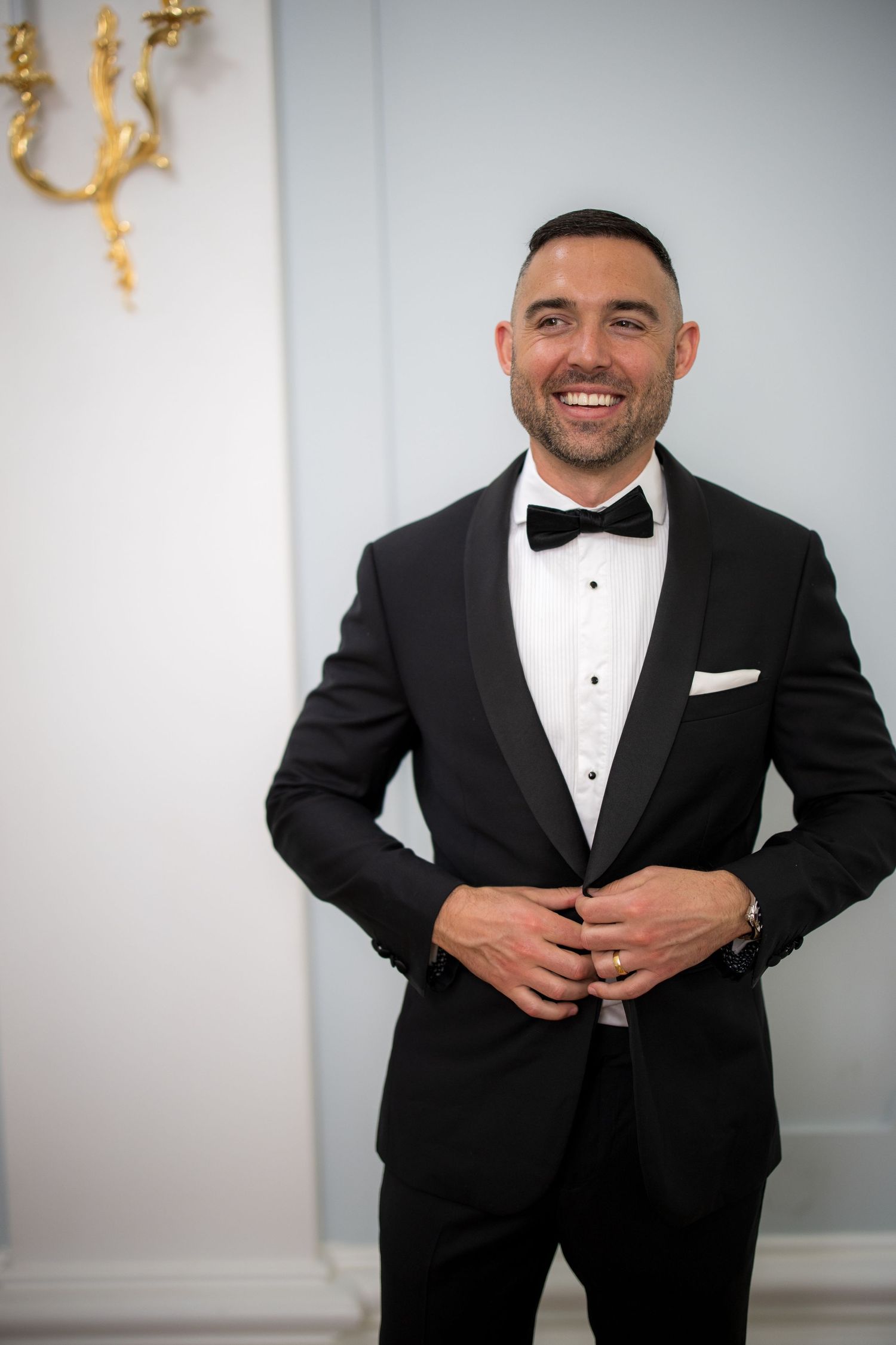 Handsome smiling groom in black tuxedo with bow tie and white pocket square poses in elegant venue.