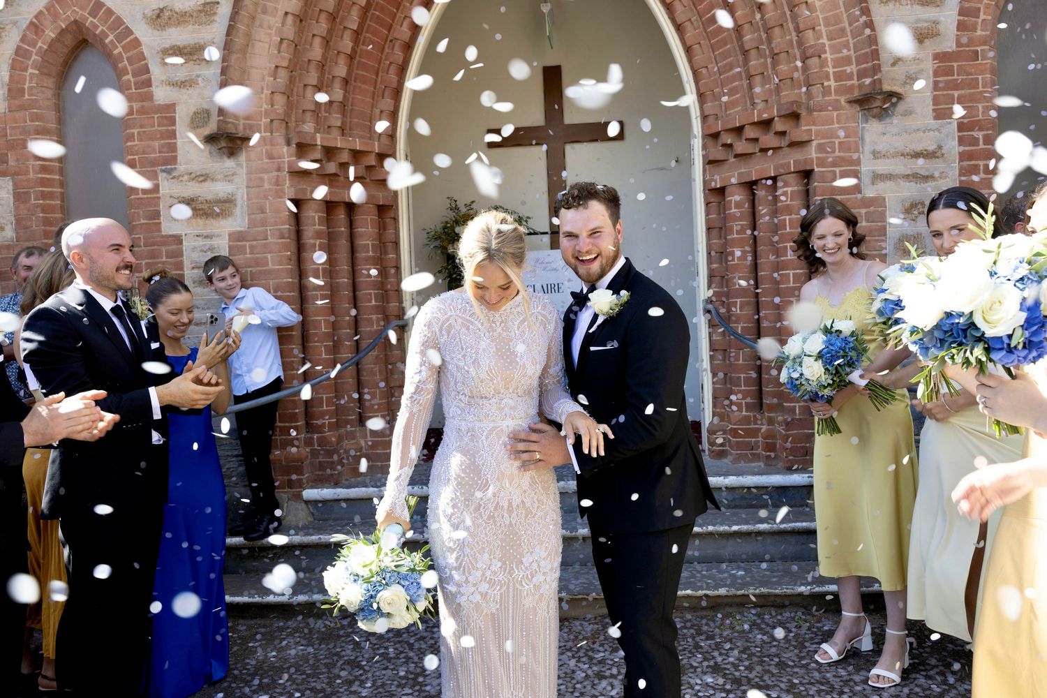 Newlyweds walking out of a brick church surrounded by guests throwing white confetti after their wedding ceremony.