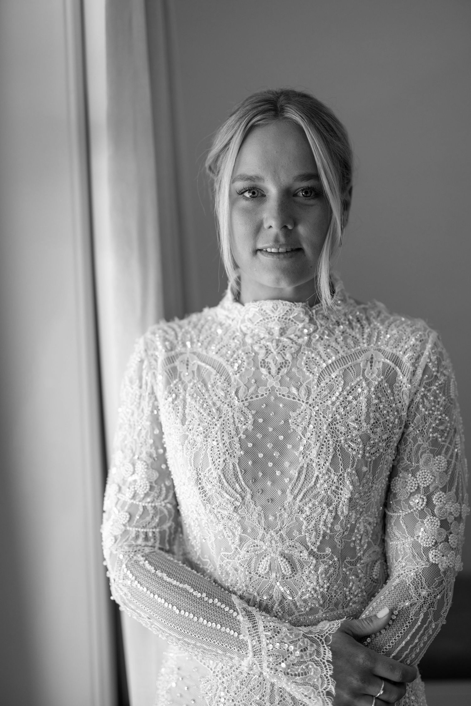 Black and white portrait of a bride in an elegant long-sleeve lace wedding gown, smiling softly near a window.