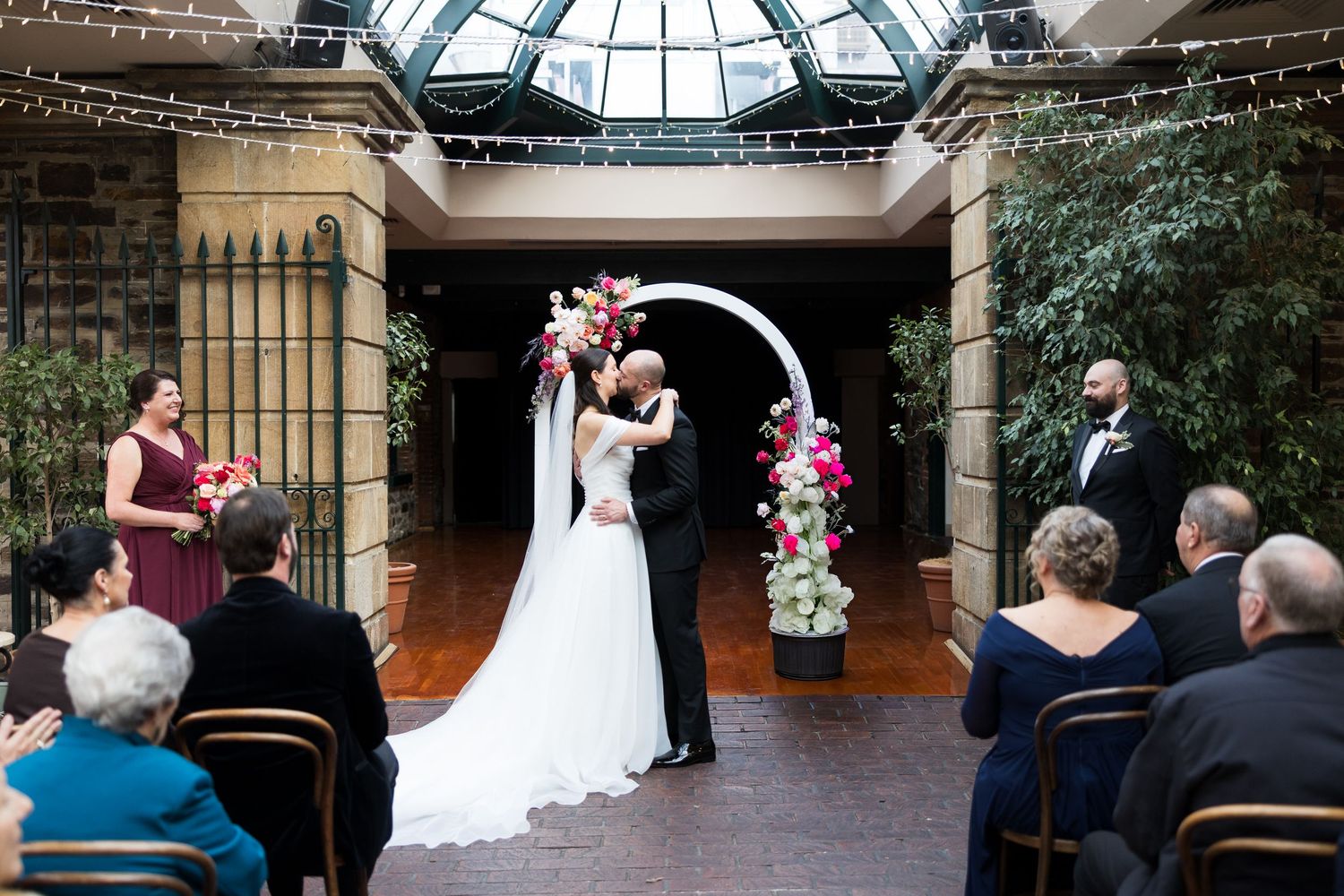 Bride and groom share first kiss under floral arch at elegant indoor wedding ceremony with seated guests.