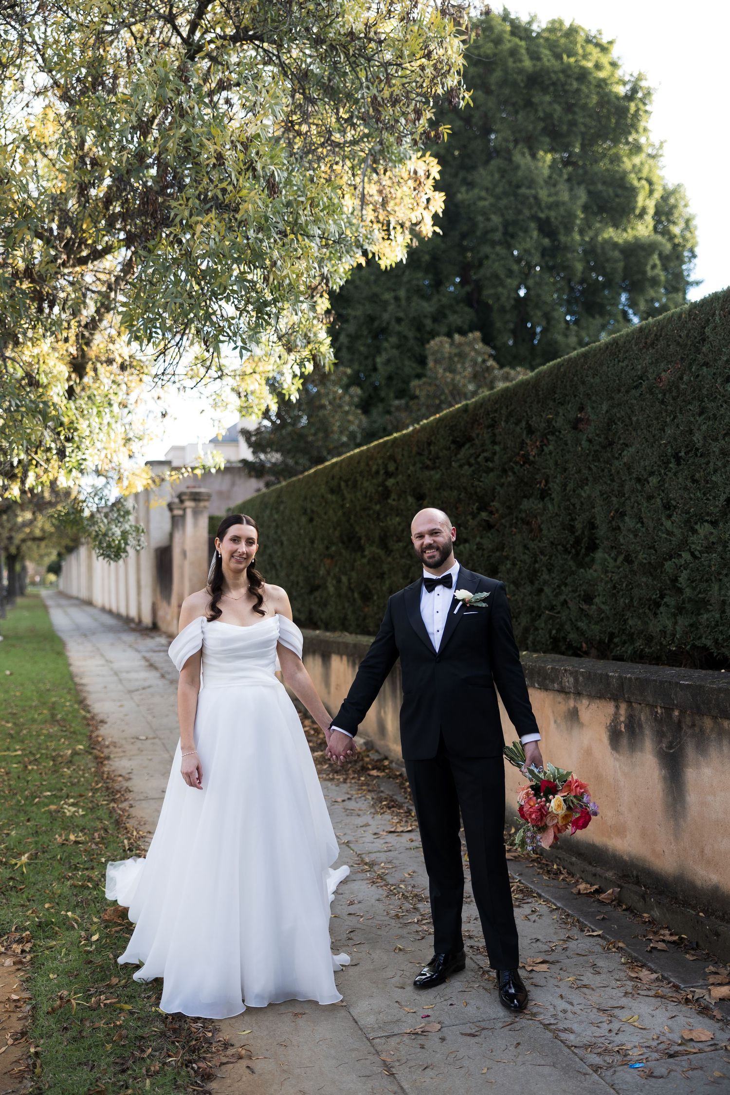 Bride and groom holding hands on a garden path, she in an off-shoulder white gown, he in a black tuxedo.
