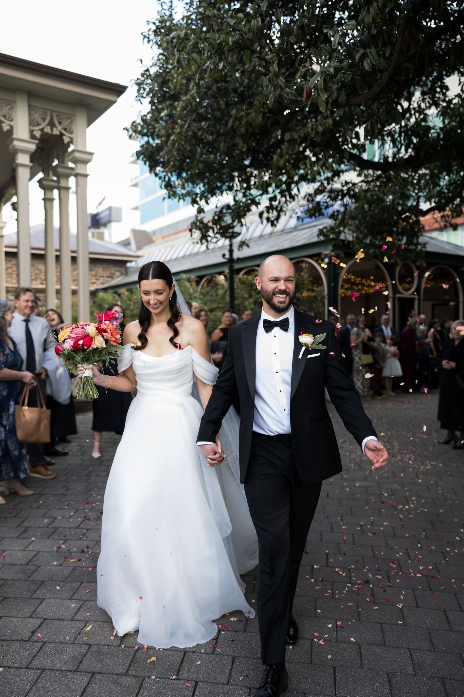 Bride and groom walk through confetti shower, she holds colorful bouquet, he wears black tuxedo, guests line outdoor venue path.