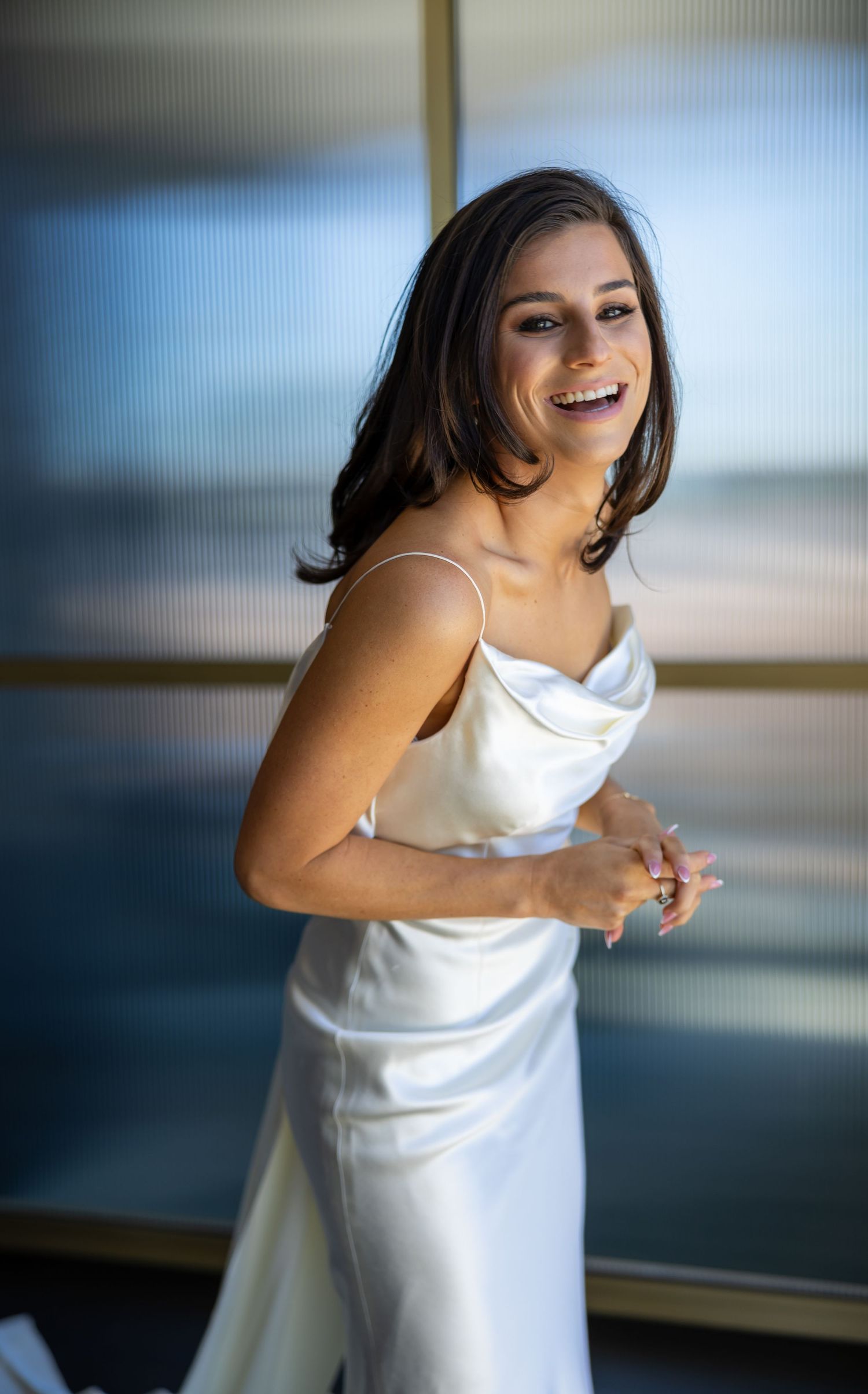 Smiling bride in elegant satin slip wedding dress laughing joyfully in soft natural light.